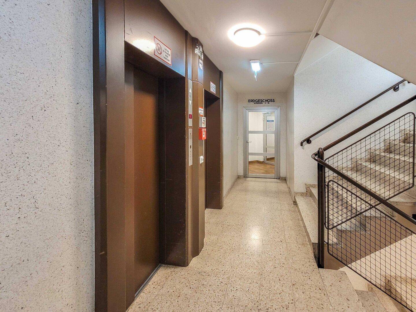 Heller Flur mit Aufzügen und Treppenhaus, führt zu den Wohneinheiten.