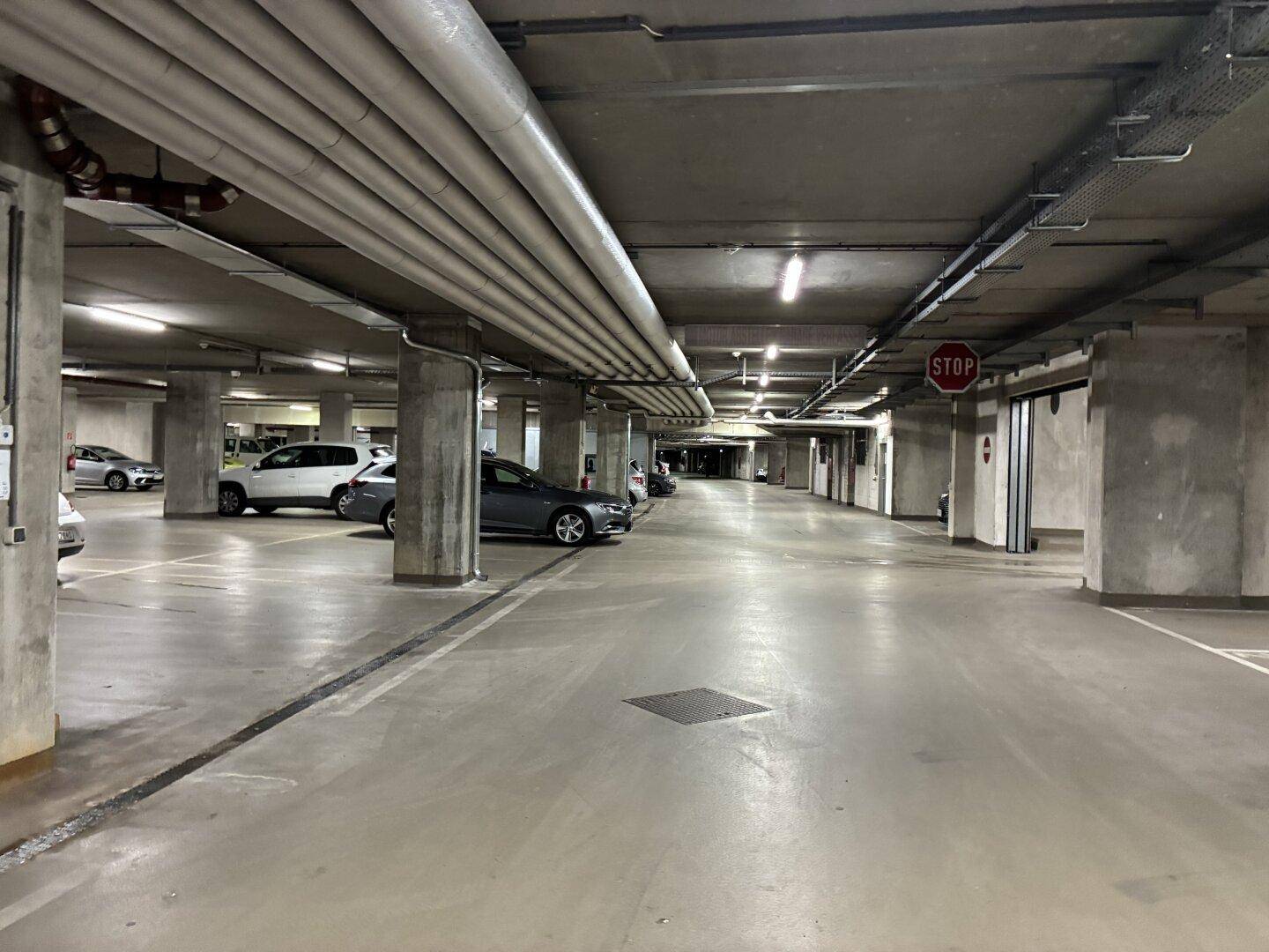 Weitläufige Tiefgarage mit Betonpfeilern, sichtbaren Rohren und markierten Stellplätzen.