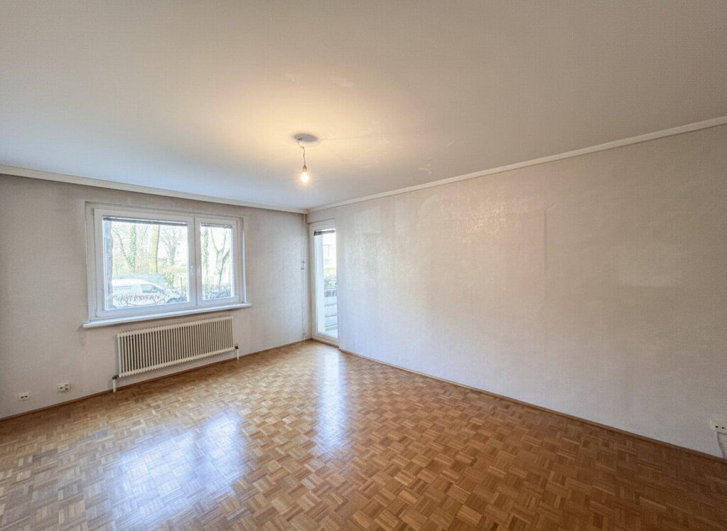 Geräumiges, leeres Wohnzimmer mit Parkettboden und großem Fenster, bereit für individuelle Gestaltung.