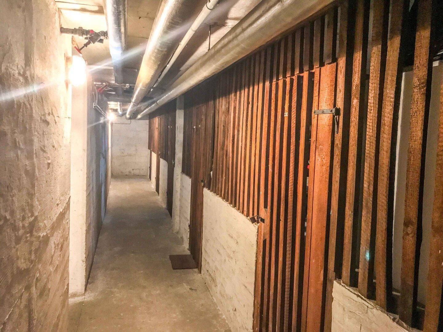 Langer Kellerflur mit individuellen Lagerabteilen und sichtbaren Rohren an der Decke.