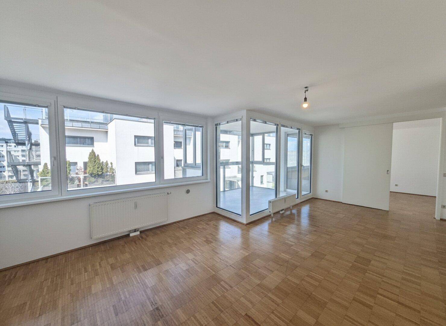 Geräumiges, leeres Wohnzimmer mit Parkettboden und großen Fenstern, die viel Tageslicht hereinlassen.