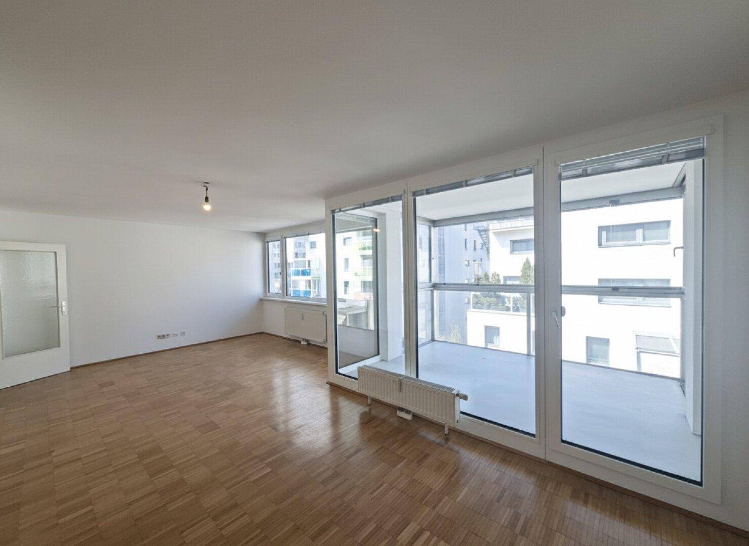Heller Wohnbereich mit Parkettboden und Zugang zu einem verglasten Balkon, der viel Licht spendet.