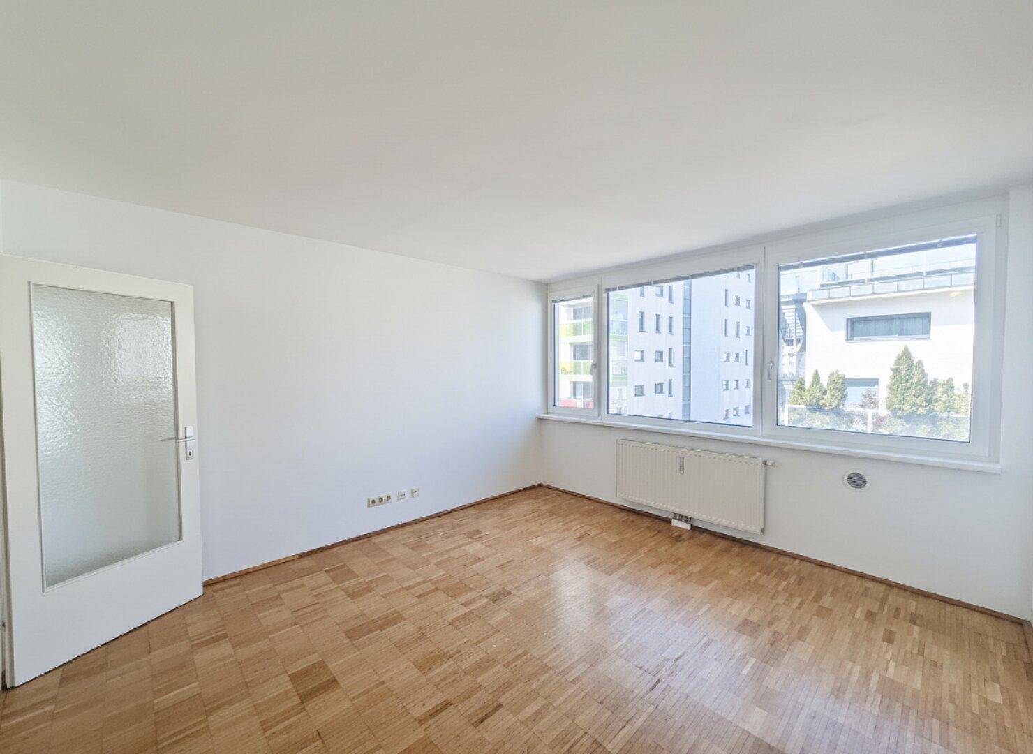 Helles Zimmer mit Parkettboden und großen Fenstern, die einen Blick auf die Nachbargebäude bieten.