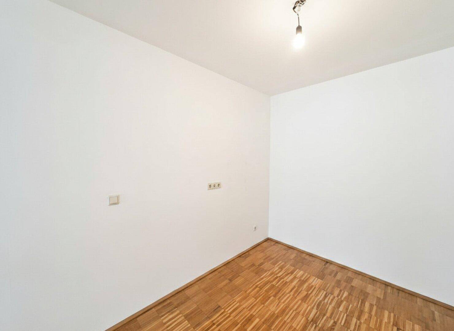 Leere Ecke eines Raumes mit Parkettboden und weißen Wänden, bereit zur Nutzung.