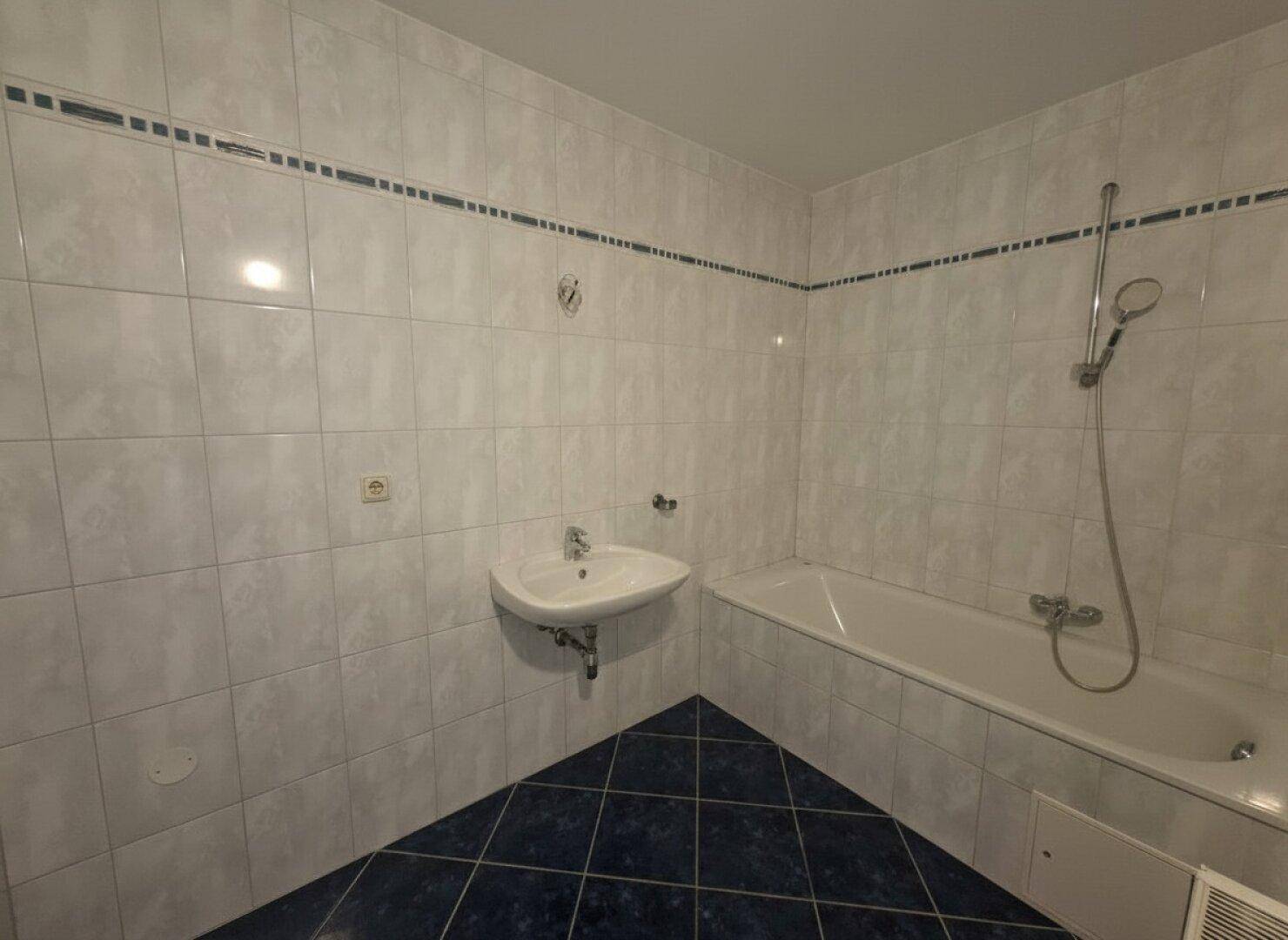 Badezimmer mit Badewanne, Waschbecken und hellen Fliesen, akzentuiert durch blaue Bodenfliesen.