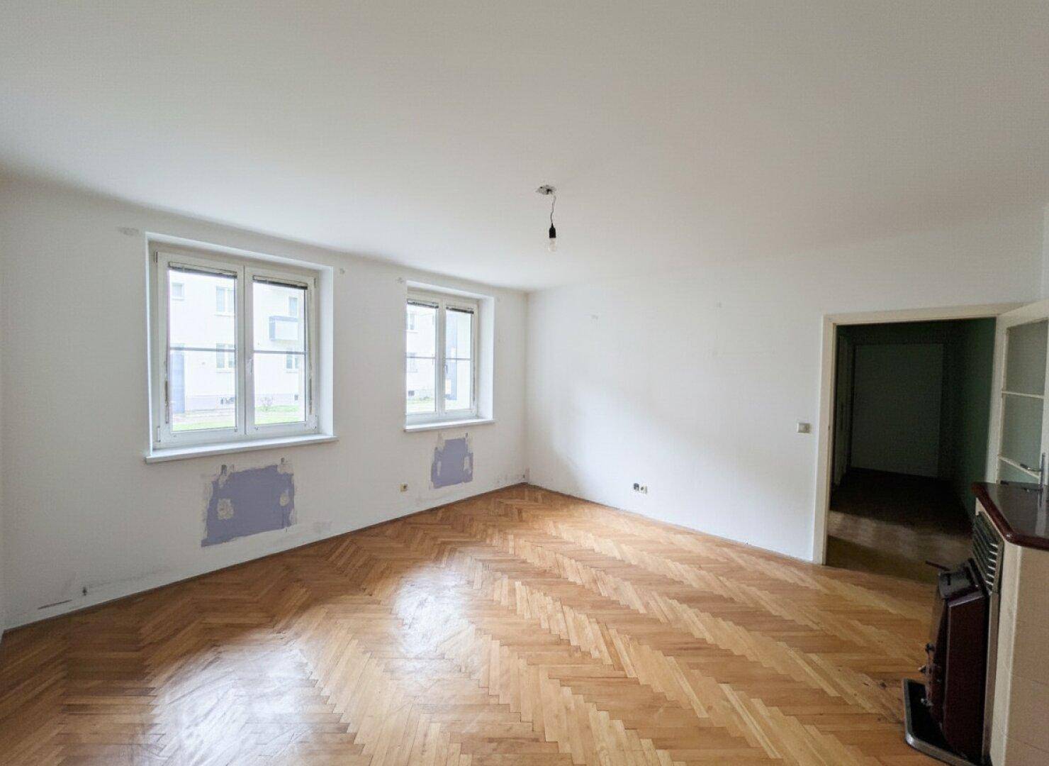 Leerer Wohnbereich mit Fischgrätparkett und zwei Fenstern, bereit zur Renovierung.