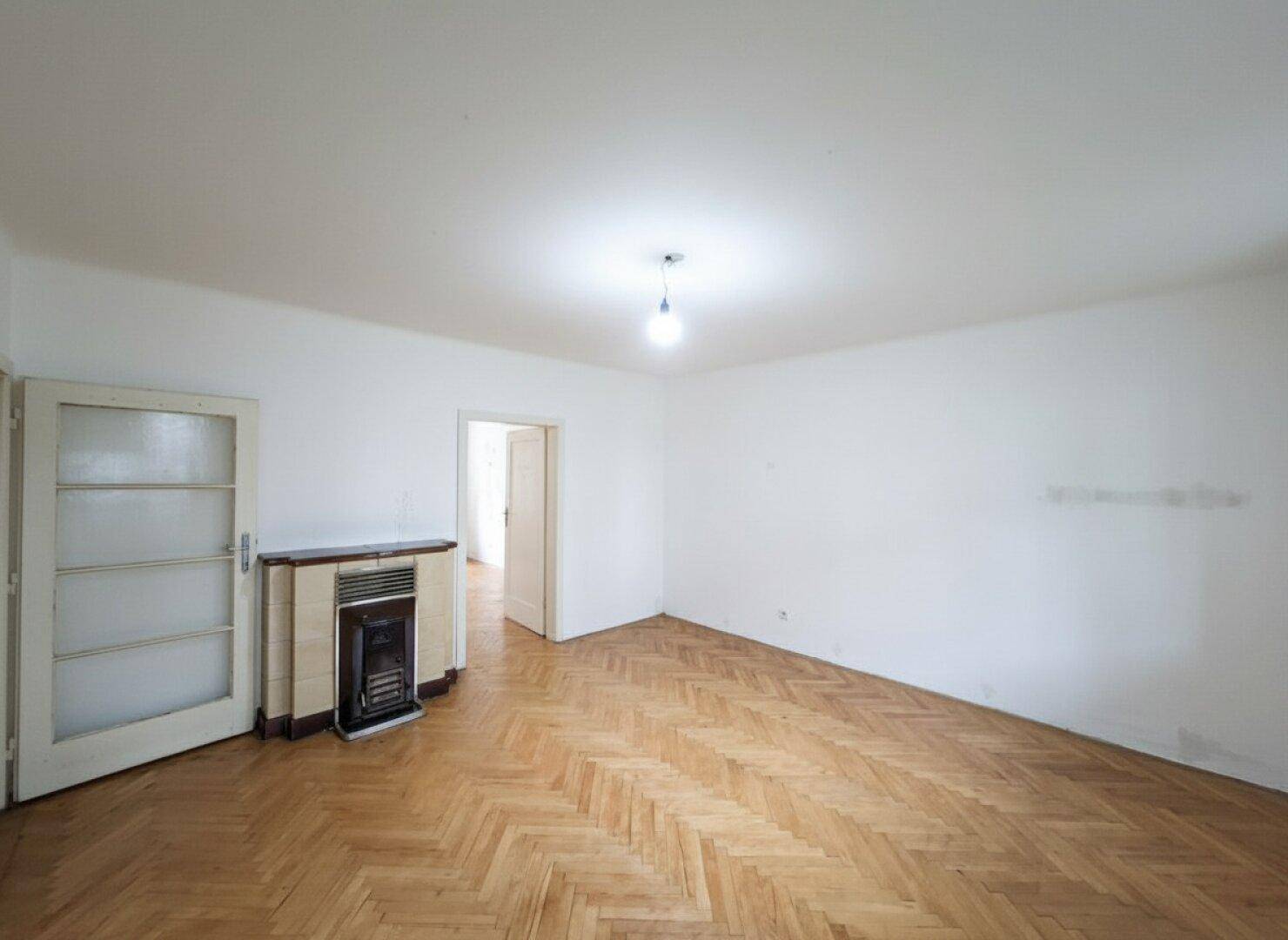 Leerer Raum mit Fischgrätparkett, einem dekorativen Ofen und einer Tür.