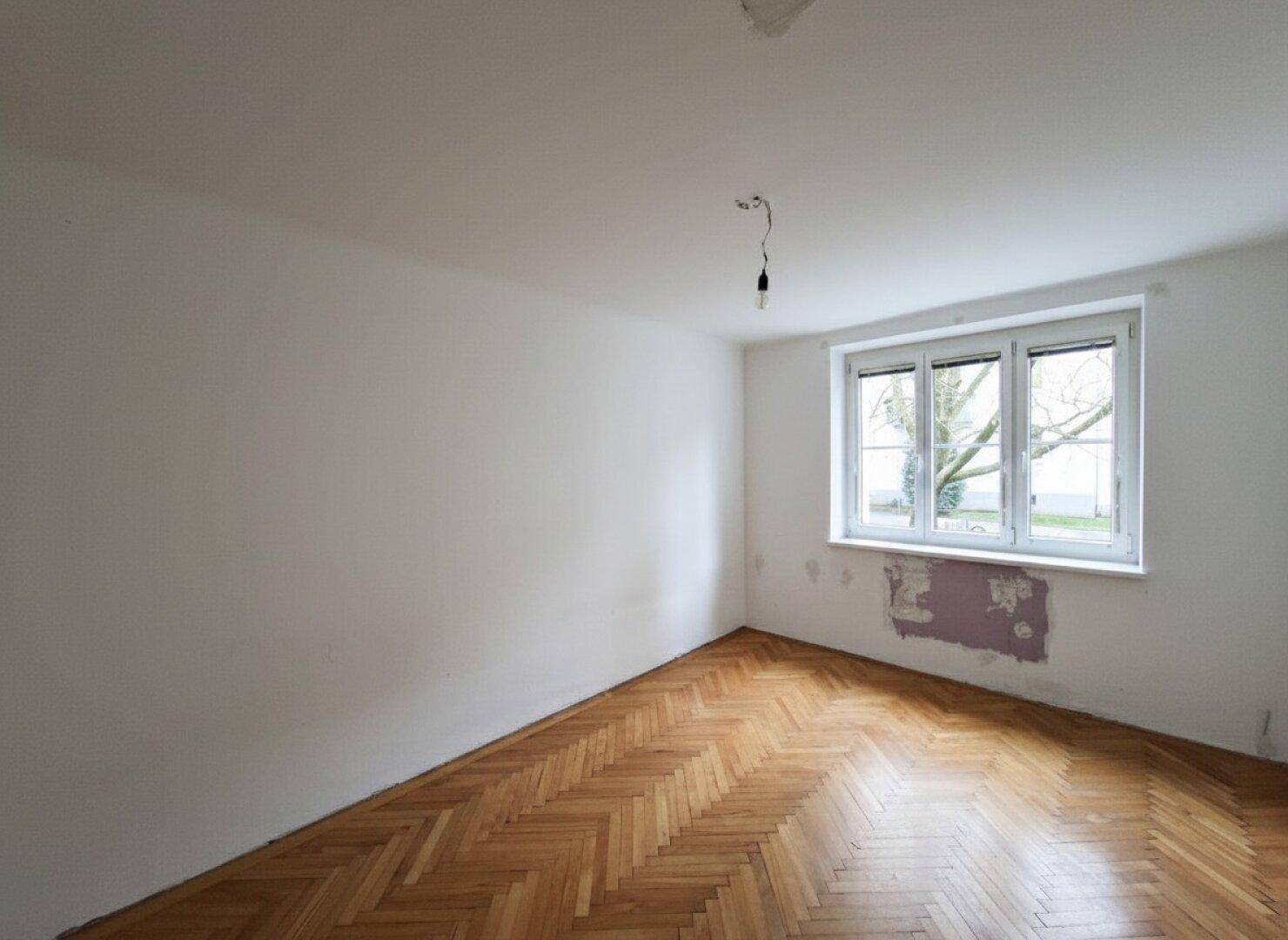 Leeres Zimmer mit Fischgrätparkett und einem großen Fenster, bereit zur Gestaltung.
