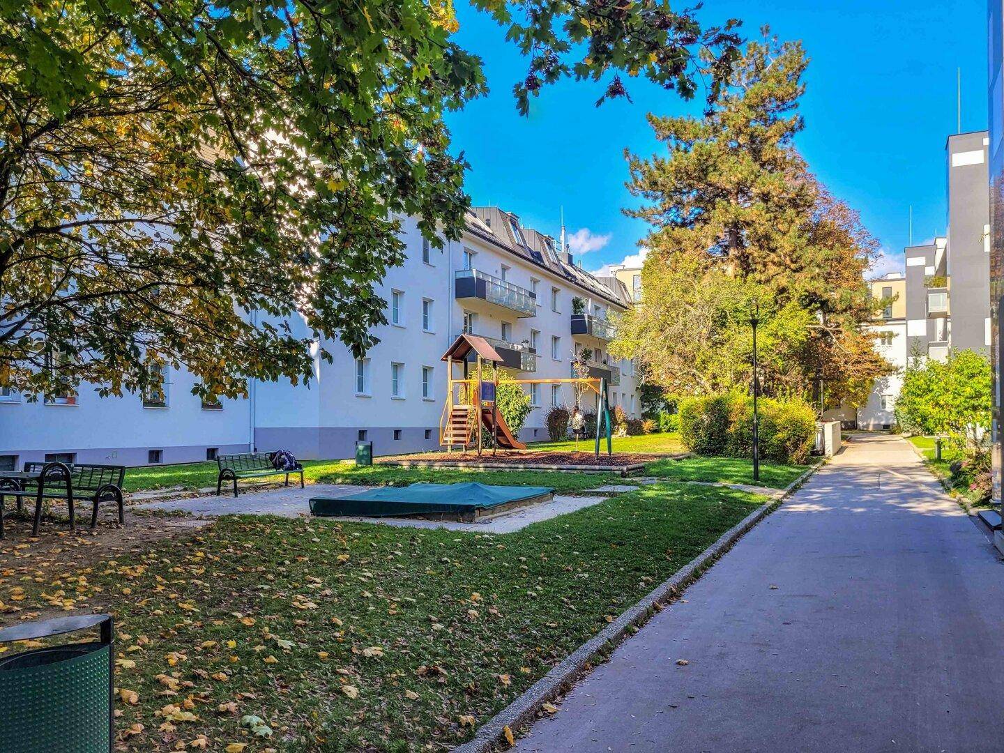 Gemeinschaftsgarten mit Spielplatz, Sandkasten und Bänken, umgeben von Bäumen.