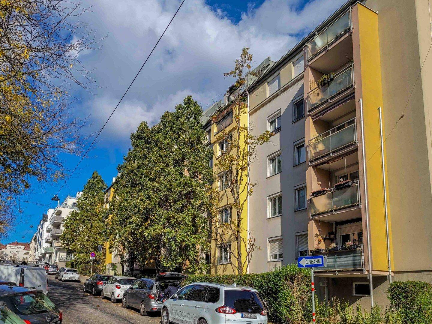 Mehrere Mehrfamilienhäuser entlang einer Straße mit parkenden Autos und Bäumen im Herbst.