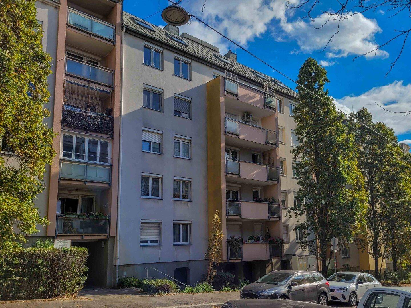 Ansicht eines Mehrfamilienhauses mit mehreren Balkonen und Bäumen unter blauem Himmel.