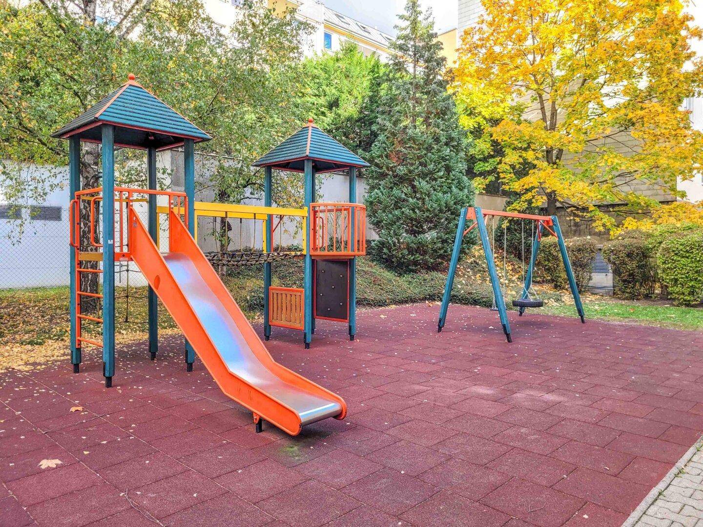 Ein bunter Spielplatz mit Rutsche und Schaukeln auf rotem Fallschutzboden, umgeben von Bäumen.
