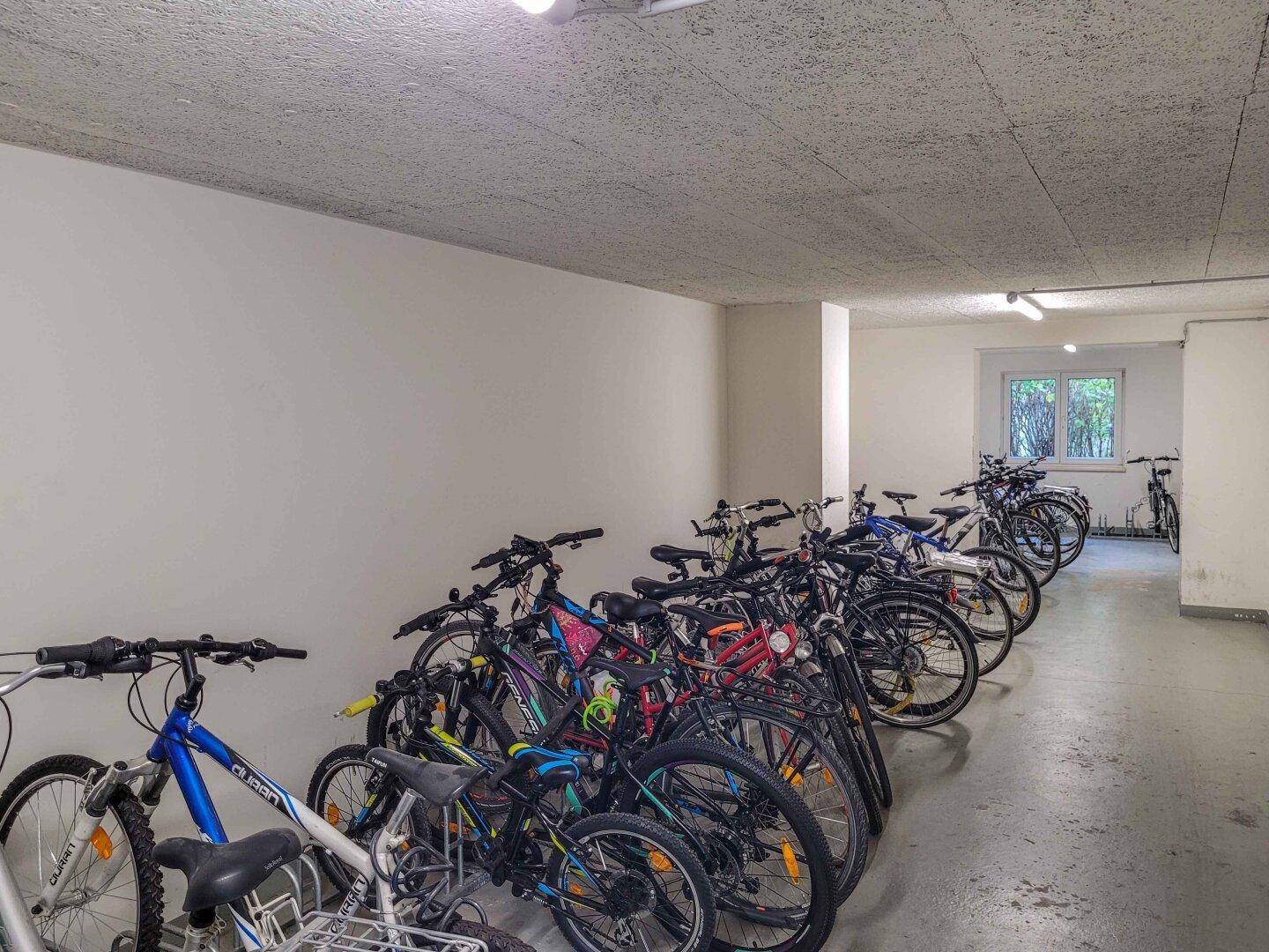 Ein heller Fahrradraum im Keller mit vielen abgestellten Fahrrädern und einem Fenster.