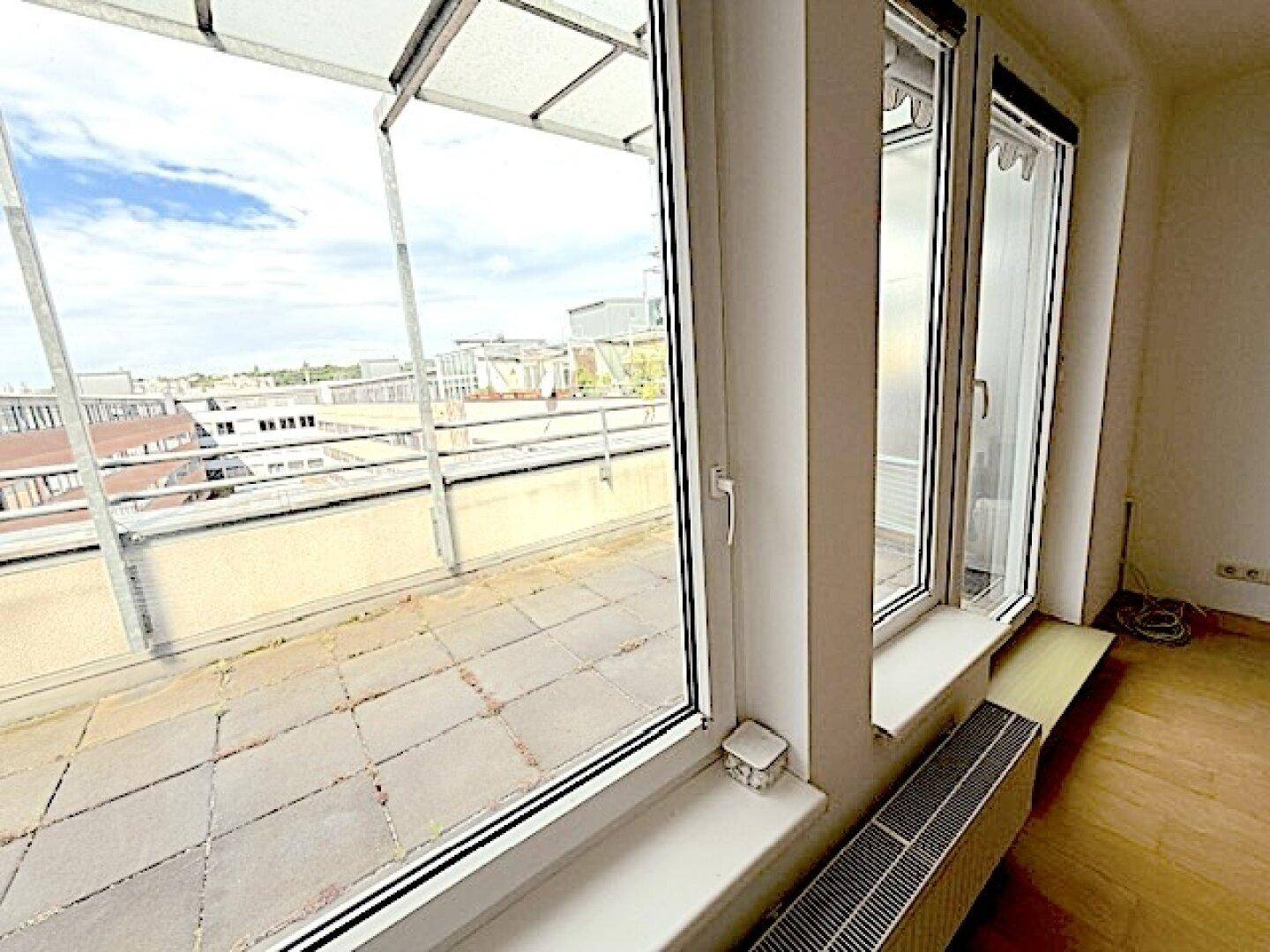 Blick aus dem Zimmer auf die geflieste Terrasse mit Überdachung und urbaner Umgebung.