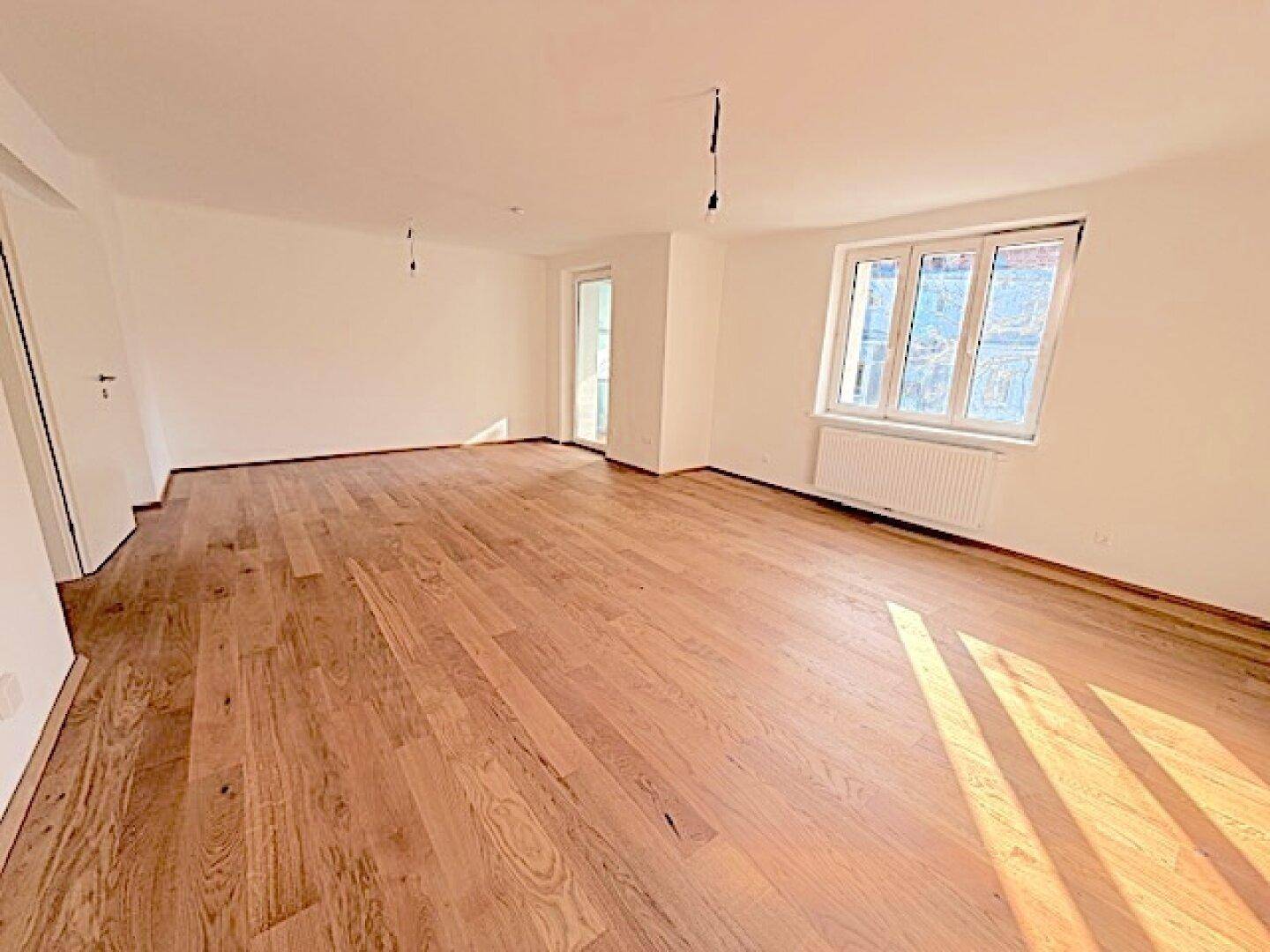 Geräumiges, leeres Zimmer mit hellem Holzboden und zwei Fenstern, bietet viel Tageslicht.