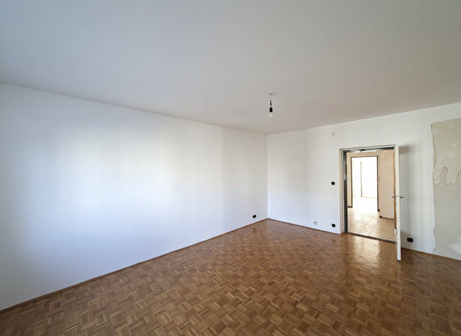 Helles, leeres Wohnzimmer mit Fischgrätparkett und weiß gestrichenen Wänden.