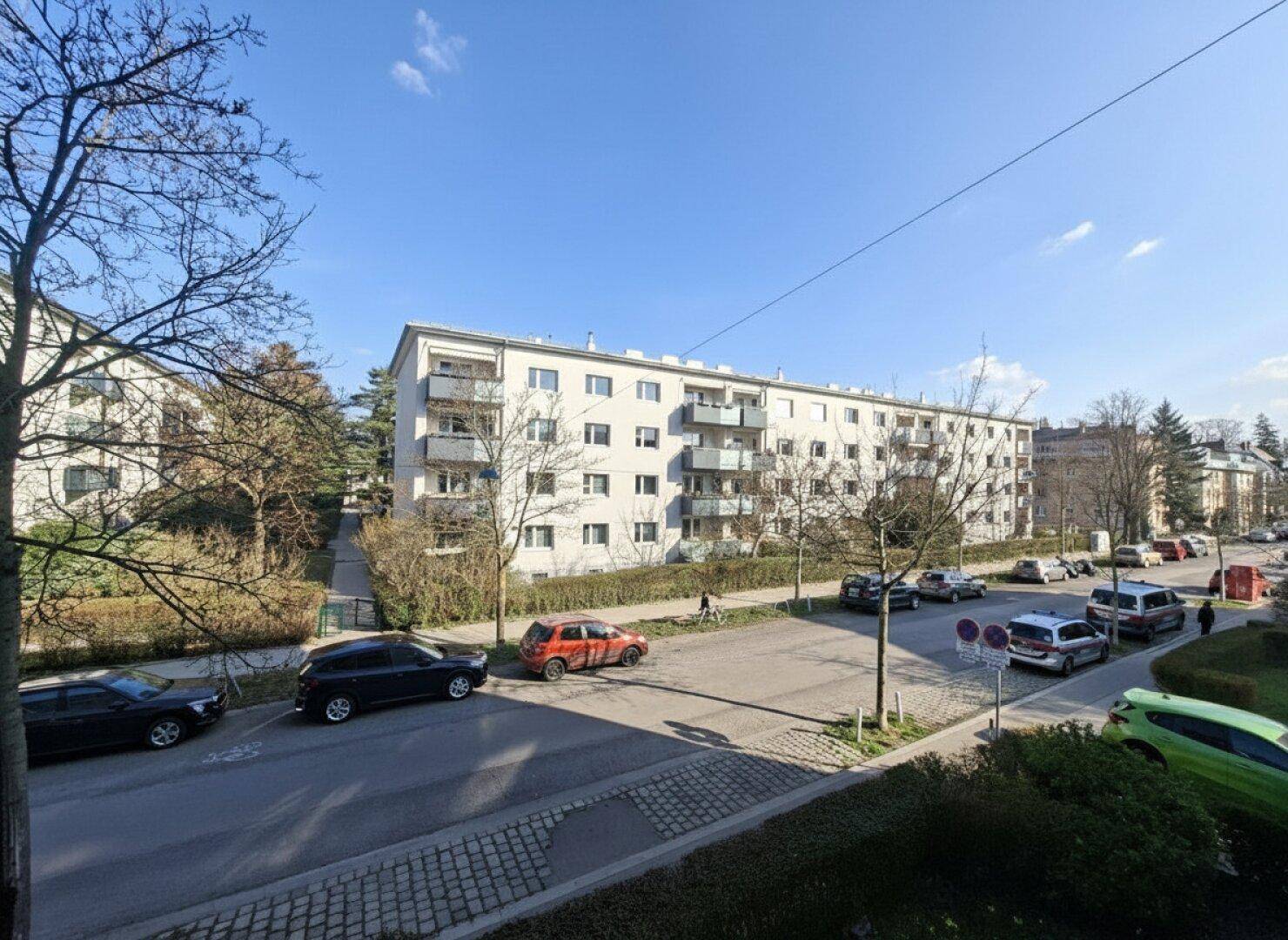 Weitläufiger Blick auf eine Wohnstraße mit einem langen Mehrfamilienhaus und viel Grün.