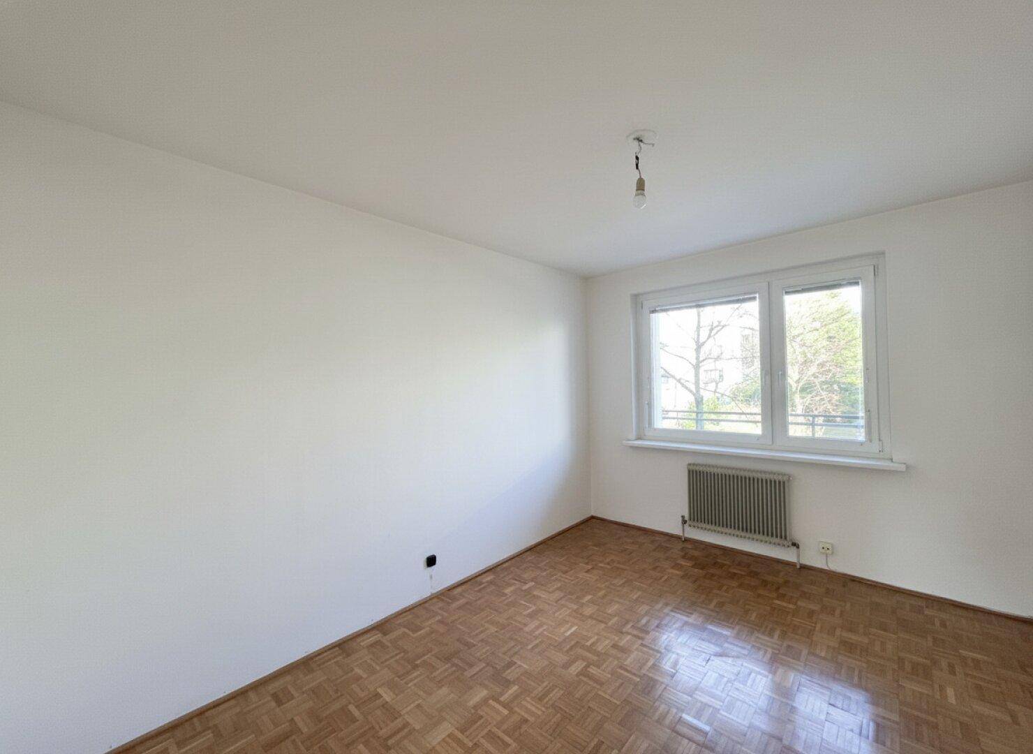 Helles, leeres Zimmer mit Parkettboden, weiß gestrichenen Wänden und einem Heizkörper unter dem Fenster.