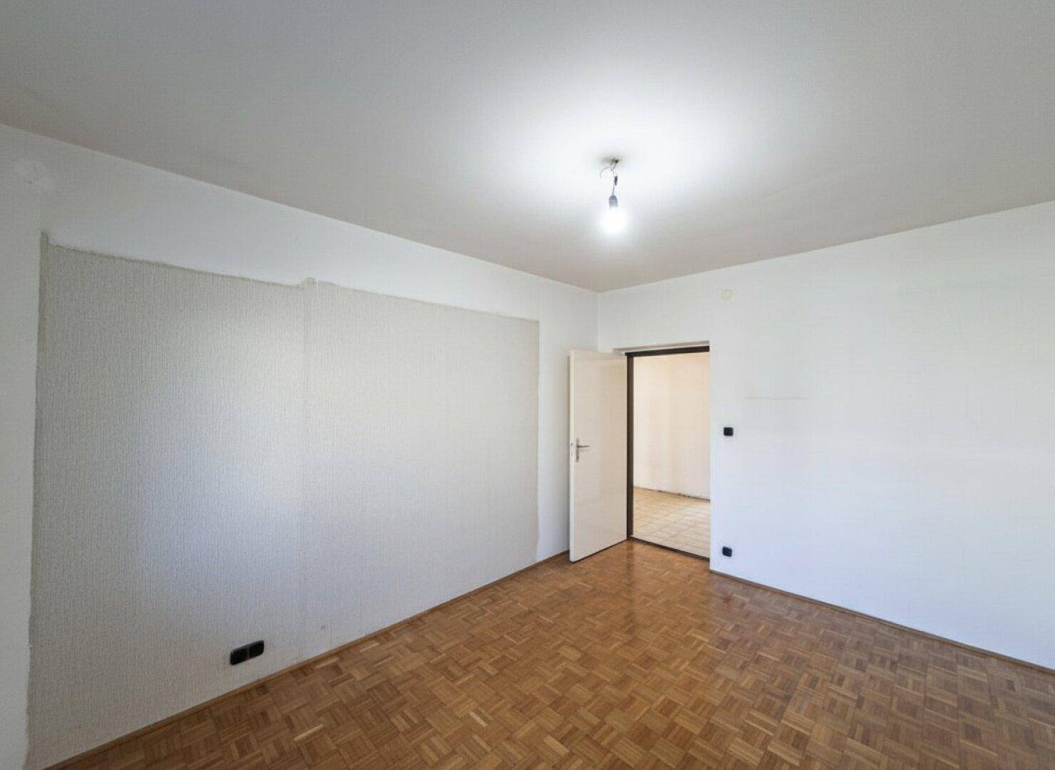 Leeres Zimmer mit Parkettboden, weißen Wänden und offener Tür zu einem weiteren Raum.