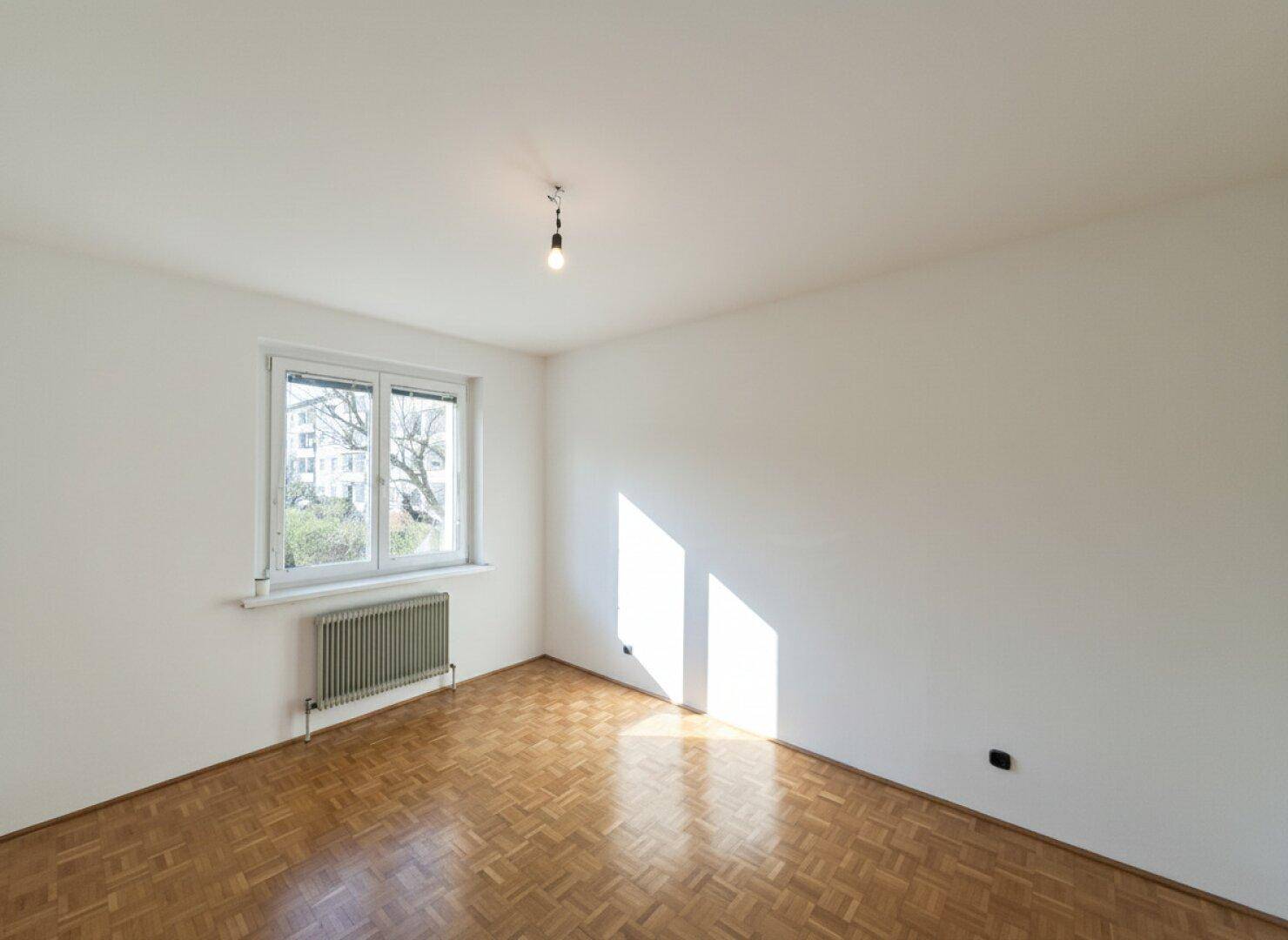 Sonniges, leeres Zimmer mit Parkettboden, weißen Wänden und einem Heizkörper unter dem Fenster.