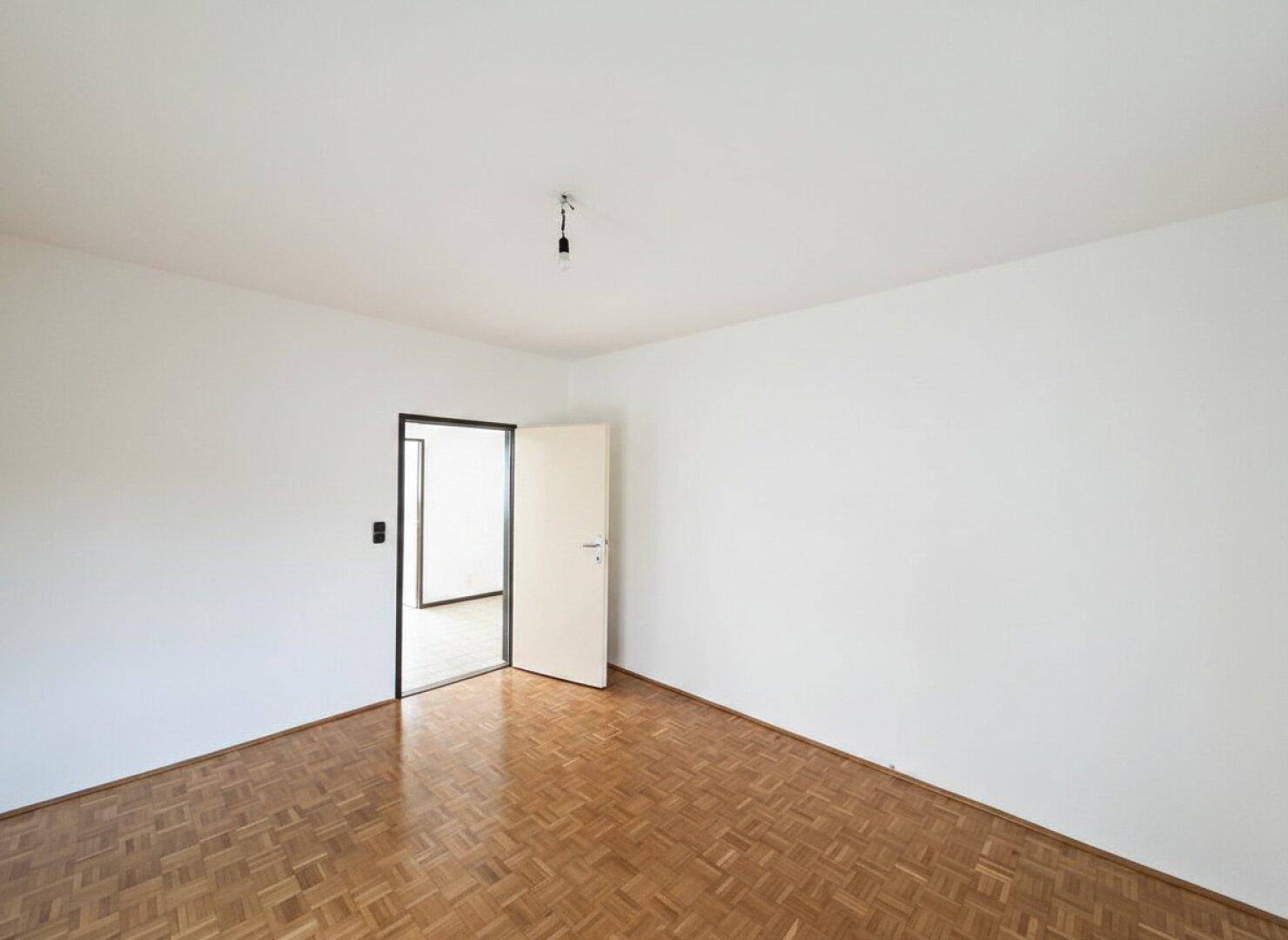 Helles, leeres Zimmer mit Parkettboden, weißen Wänden und offener Tür zu einem weiteren Bereich.