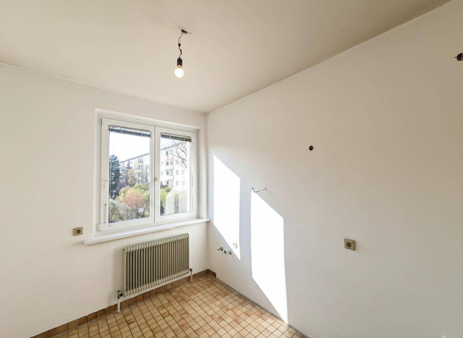 Leere Küche mit Fliesenboden, weißen Wänden, Fenster und Heizkörper, bereit für die Einrichtung.