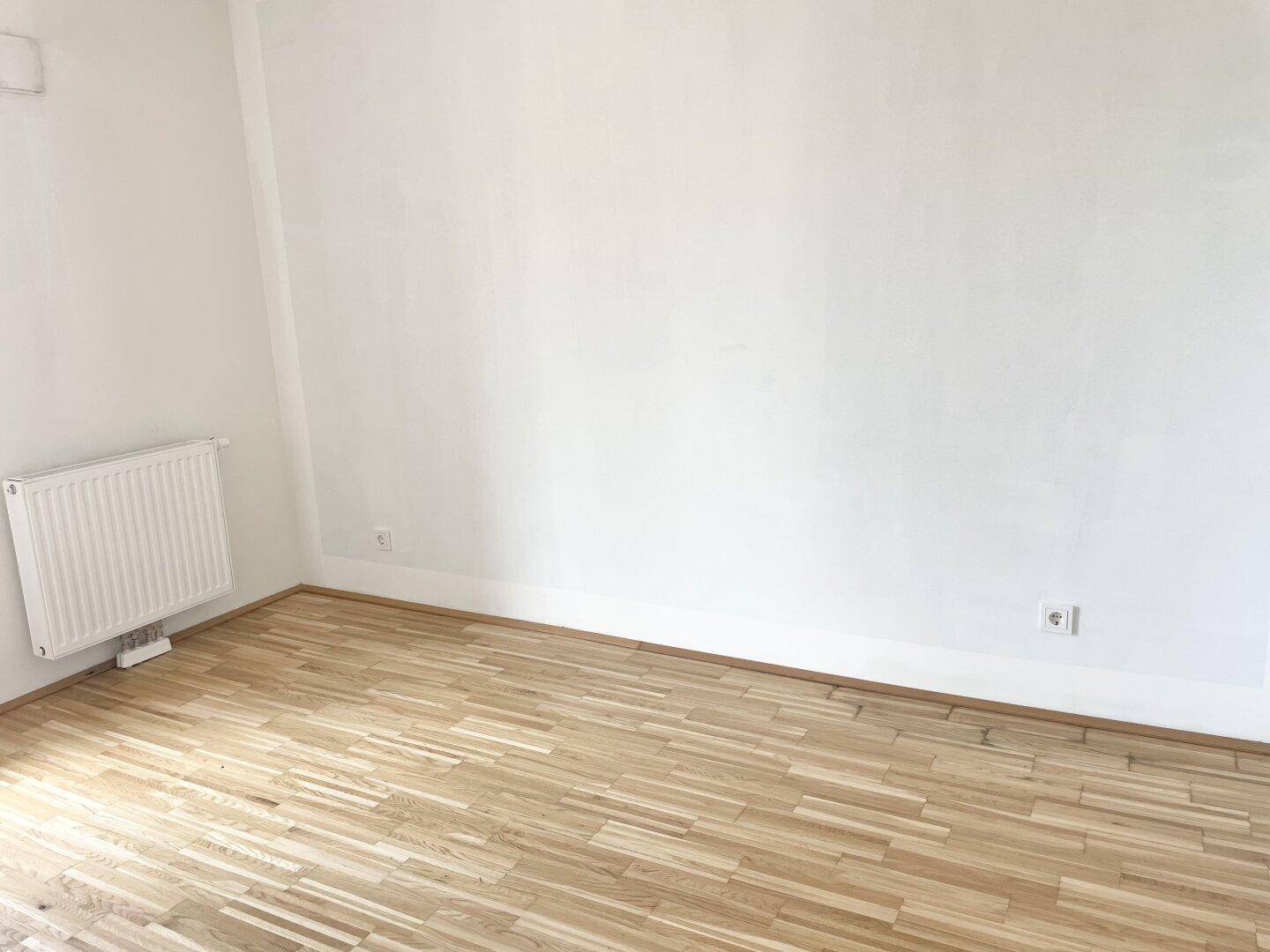 Geräumiges, helles Zimmer mit Parkettboden und weißen Wänden.