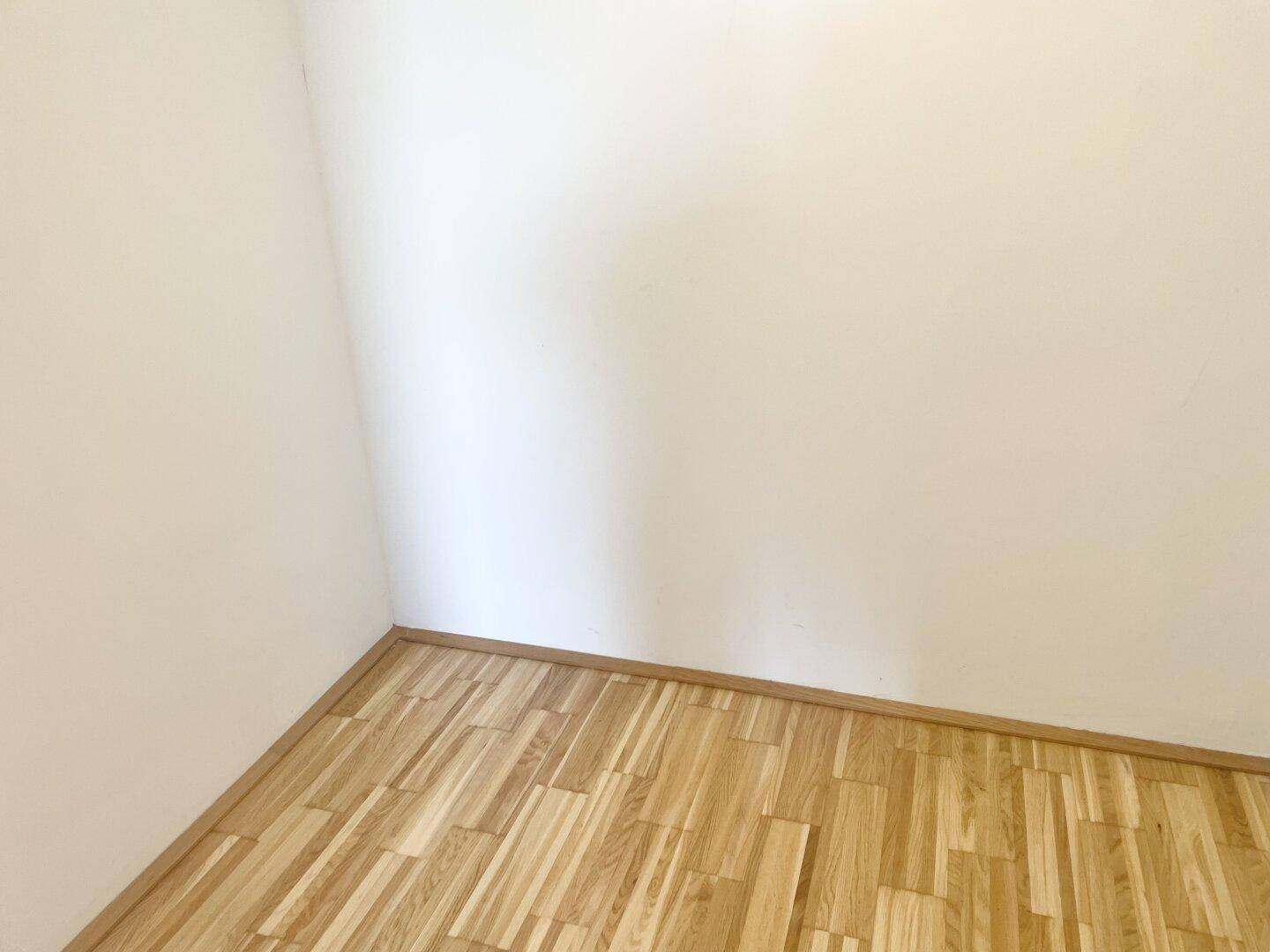Ecke eines hellen Zimmers mit Parkettboden und weißen Wänden.