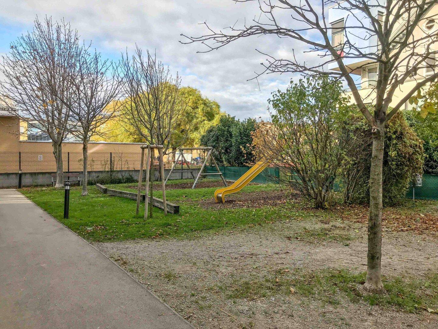Kinderspielplatz mit Rutsche und Schaukeln in einer grünen Umgebung mit Bäumen.