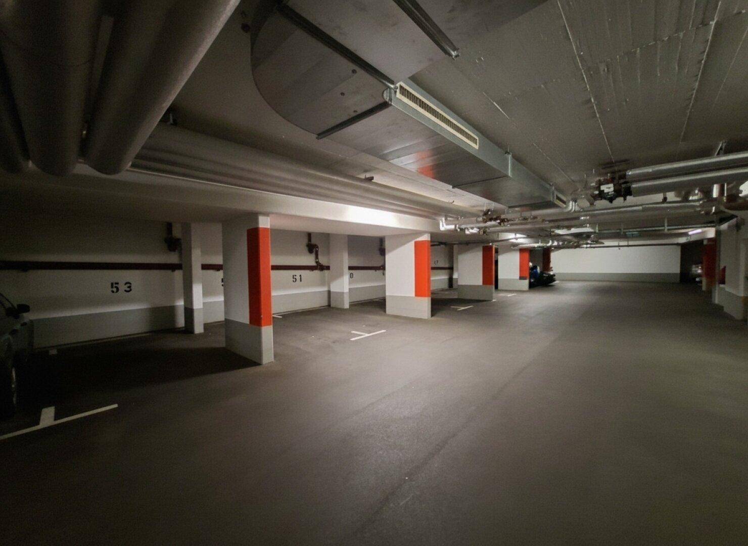 Leere Tiefgarage mit nummerierten Stellplätzen, roten Säulen und Deckeninstallationen.