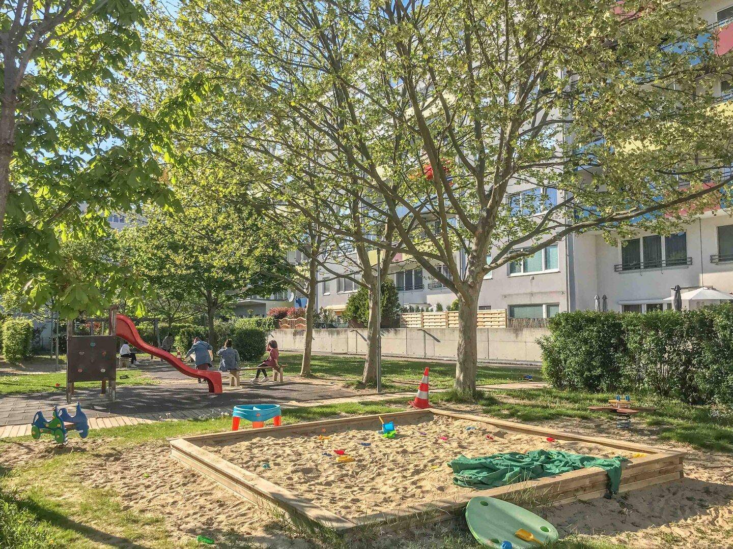 Heller Gemeinschaftsgarten mit Spielplatz, Sandkasten und Rutsche, umgeben von Bäumen.