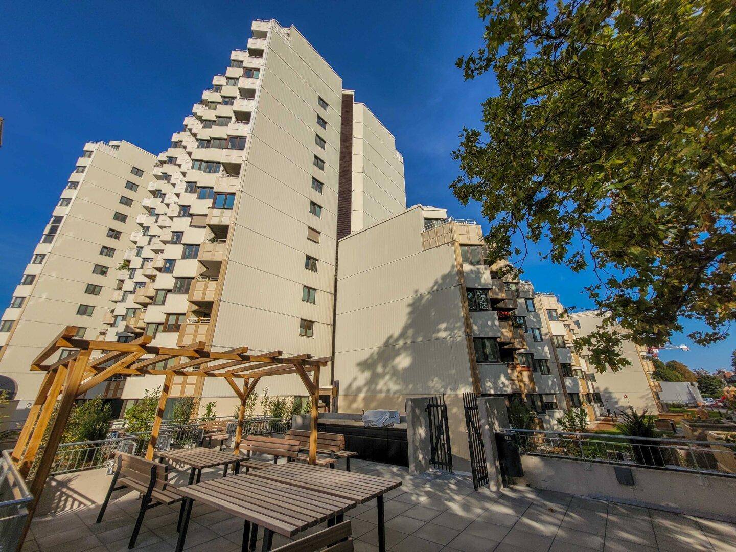 Außenbereich mit Pergola, Holztischen und Bänken vor dem Apartmentgebäude.