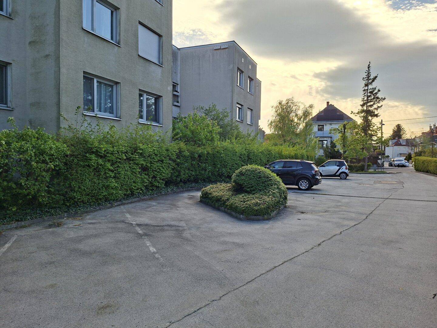 Außenansicht eines Wohnhauses mit Parkplätzen, umgeben von grünen Hecken und Bäumen.