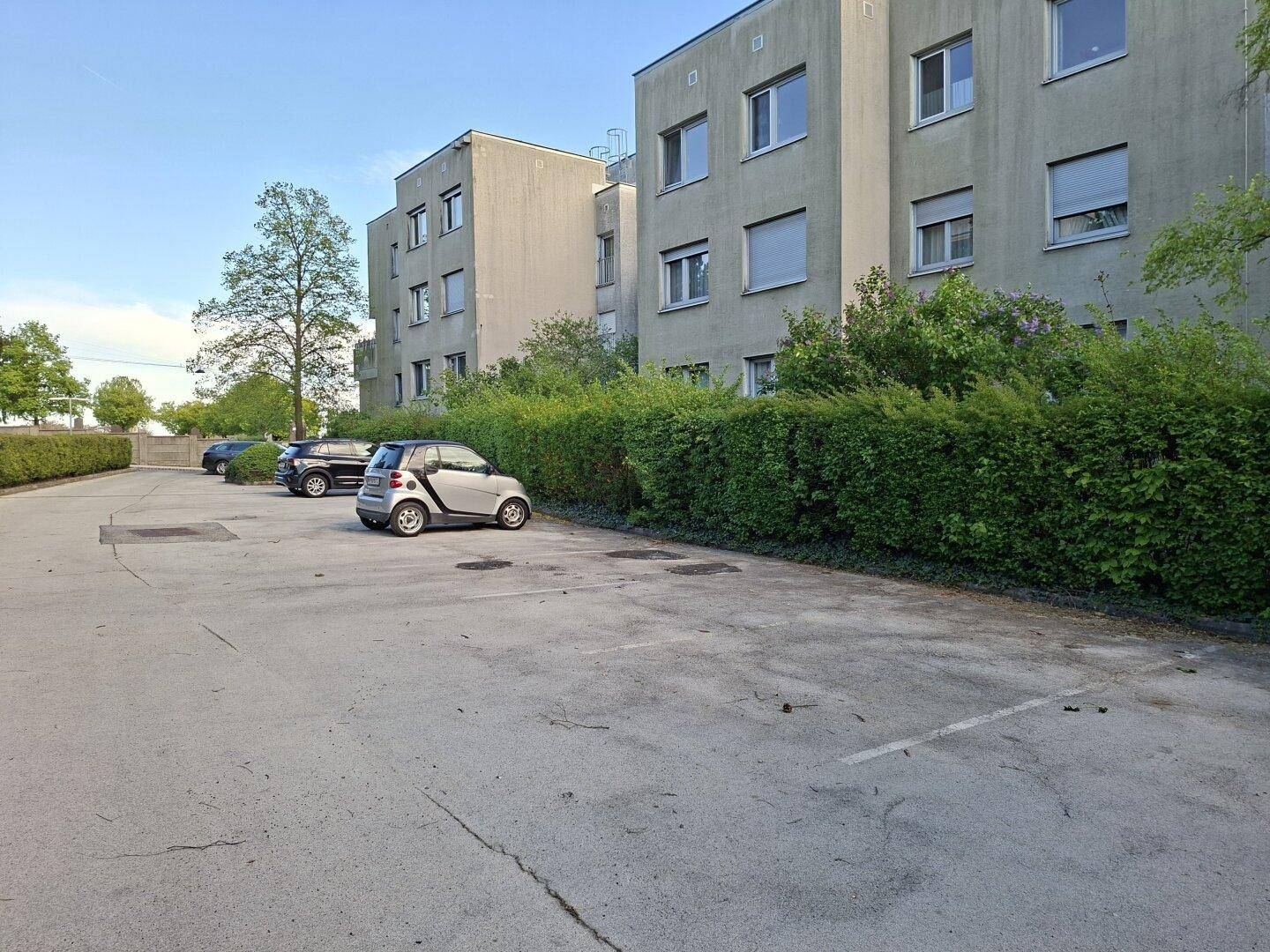 Mehrere Wohngebäude mit großem Parkplatz, begrenzt durch eine lange grüne Hecke.