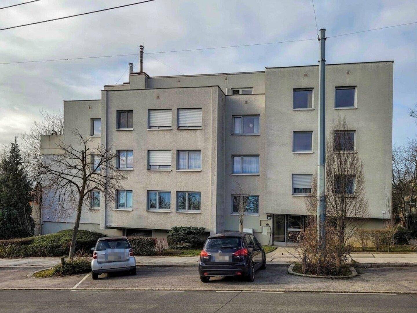 Vorderansicht eines Wohnhauses mit mehreren Etagen und geparkten Autos auf der Straße.