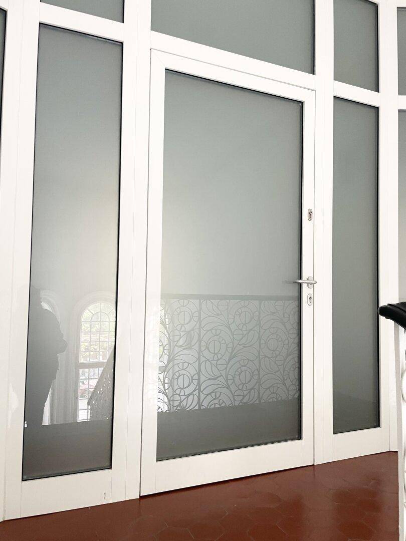 Weiße Glastür mit dekorativem Muster und einem Blick auf ein Fenster im Hintergrund.
