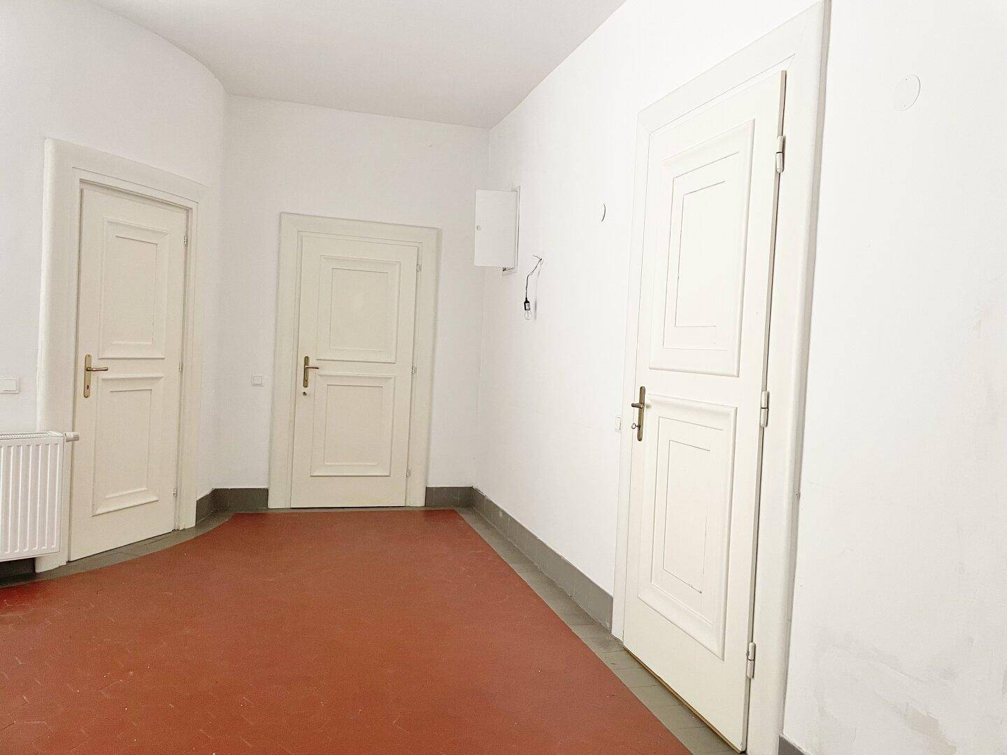 Geräumiger Flur mit drei weißen Türen, hellen Wänden und einem rotbraunen Fliesenboden.