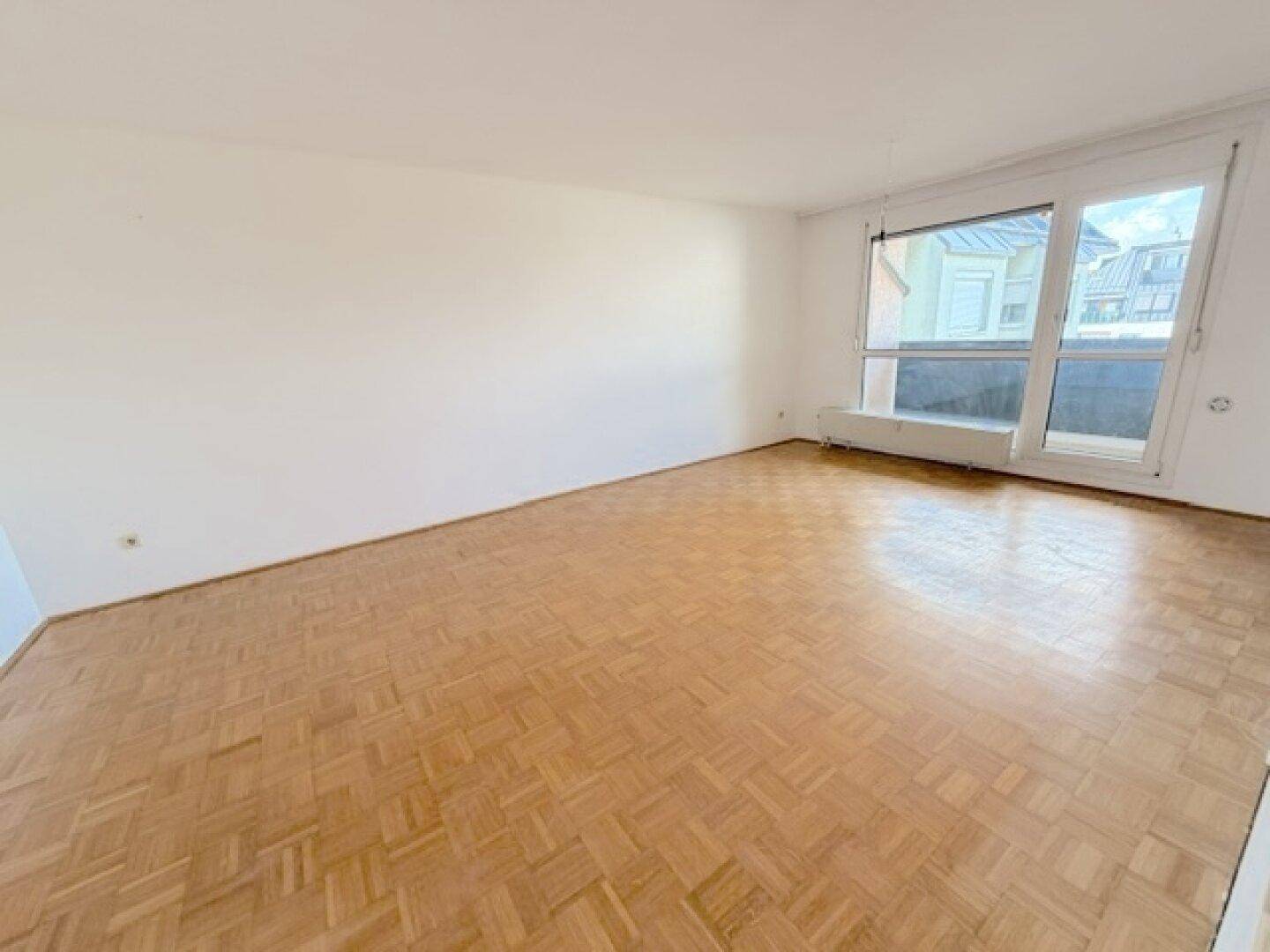 Leeres Wohnzimmer mit Fischgrätparkett und Zugang zu einem Balkon, bietet viel natürliches Licht.