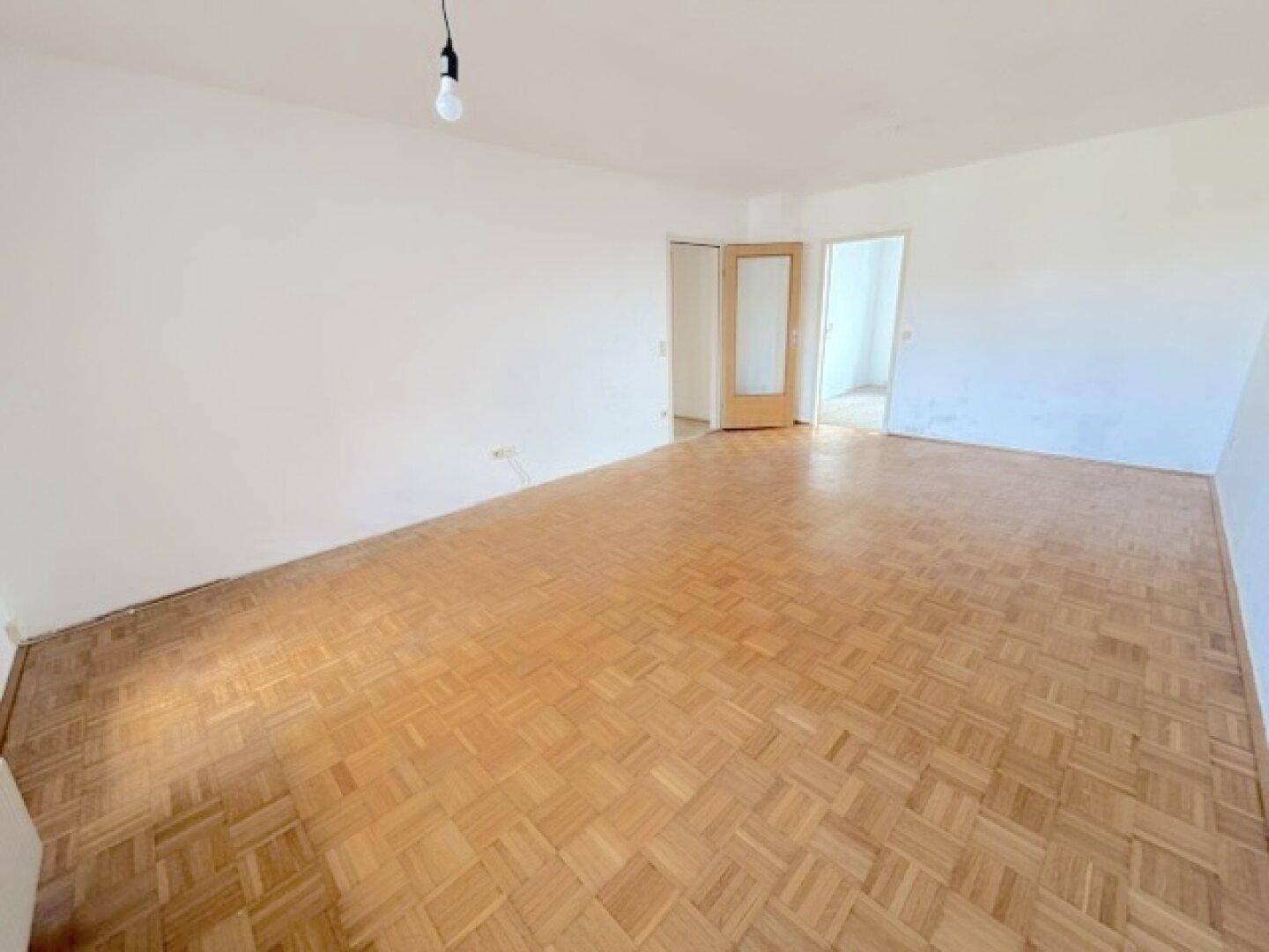 Geräumiges, leeres Wohnzimmer mit Fischgrätparkett und Zugang zu weiteren Räumen.