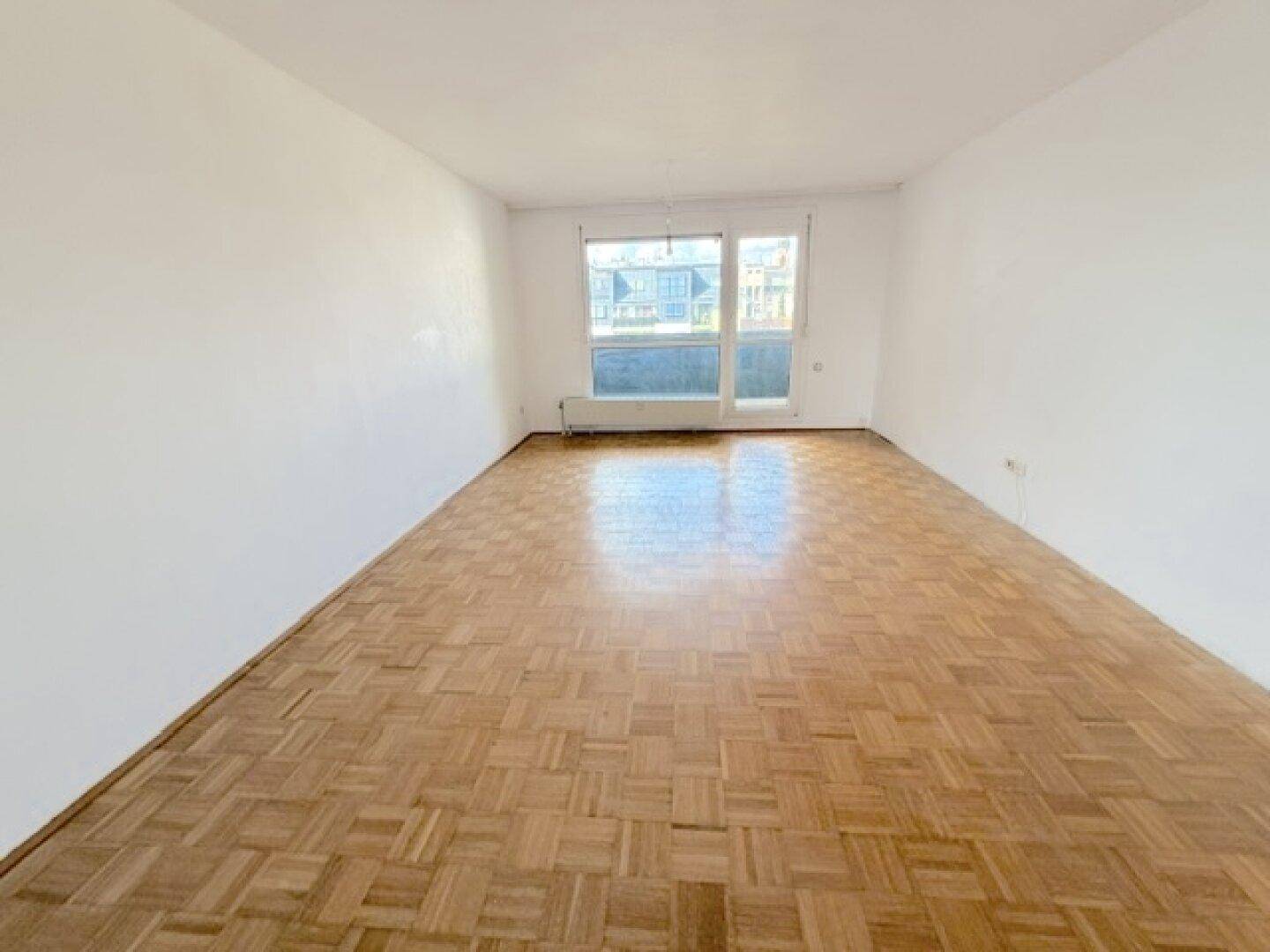 Geräumiges Wohnzimmer mit Fischgrätparkett und großem Fenster, bietet viel Helligkeit.
