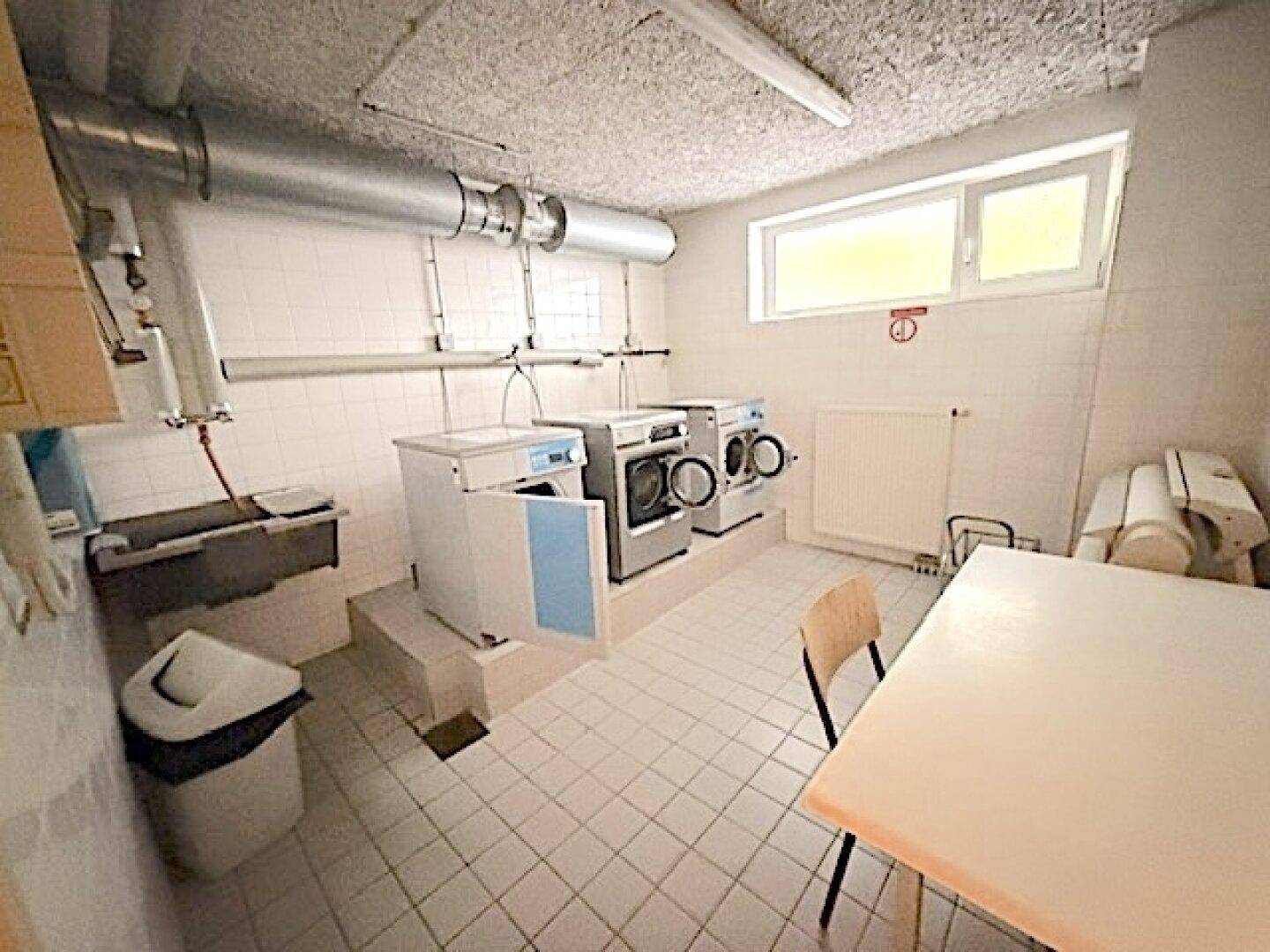 Gemeinschaftlicher Waschraum mit Waschmaschinen, Spülbecken und Fenster.
