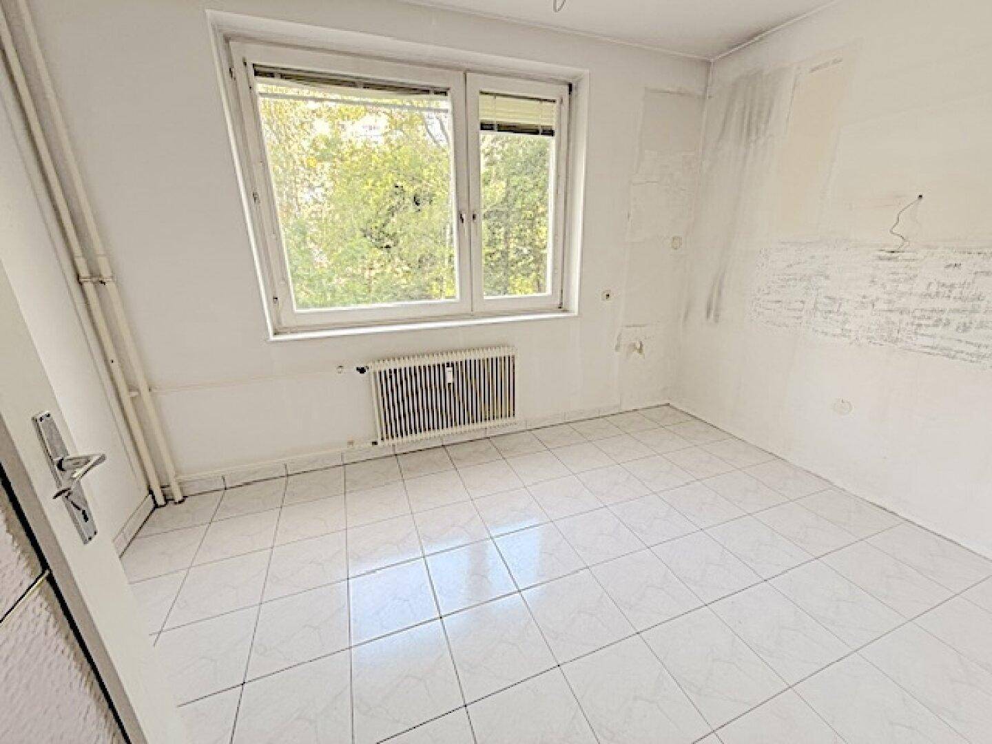 Leeres Zimmer mit weißen Fliesen, großem Fenster und Blick ins Grüne, bereit zur individuellen Gestaltung.