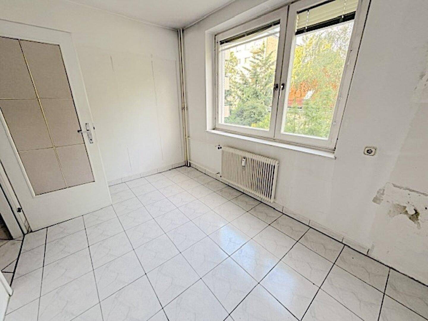 Helles Zimmer mit Fliesenboden und großem Fenster, das viel Tageslicht hereinlässt und eine grüne Aussicht bietet.