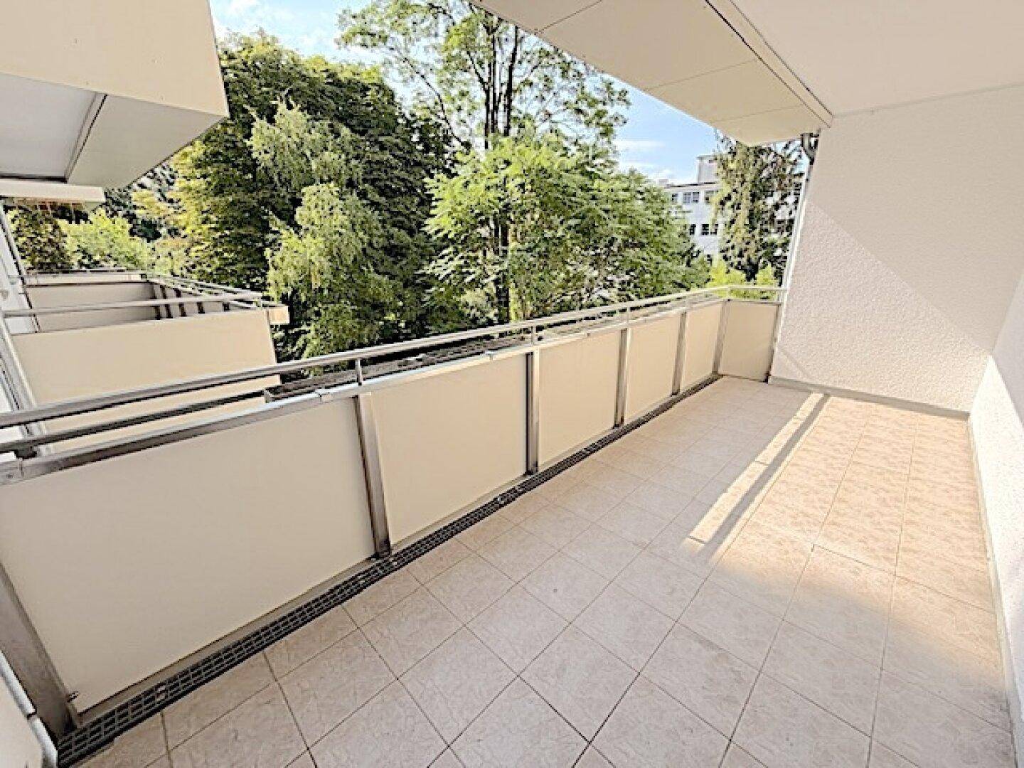 Heller Balkon mit Fliesenboden und weitem Blick auf die grüne Umgebung und den Himmel.
