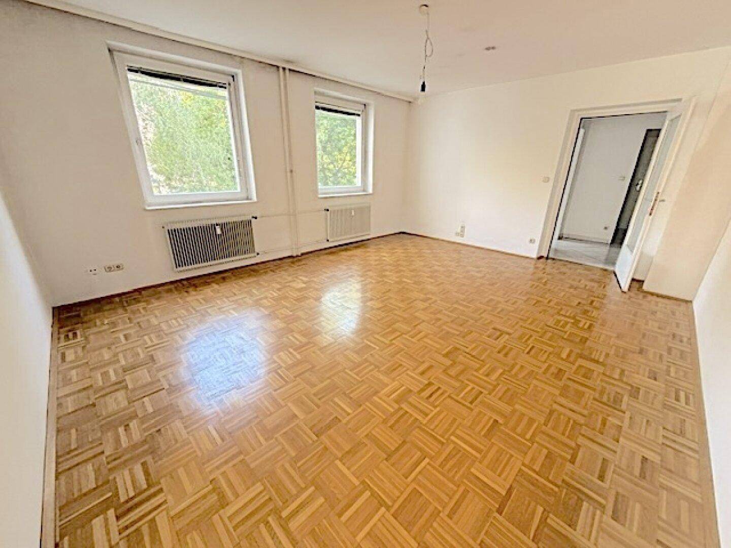 Helles, leeres Wohnzimmer mit Fischgrätparkett und zwei großen Fenstern mit Blick ins Grüne.