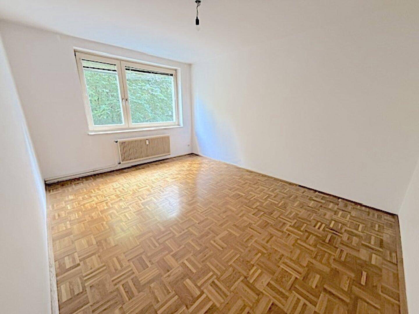 Geräumiges Zimmer mit Fischgrätparkett und großem Fenster, das viel Tageslicht und eine grüne Aussicht bietet.