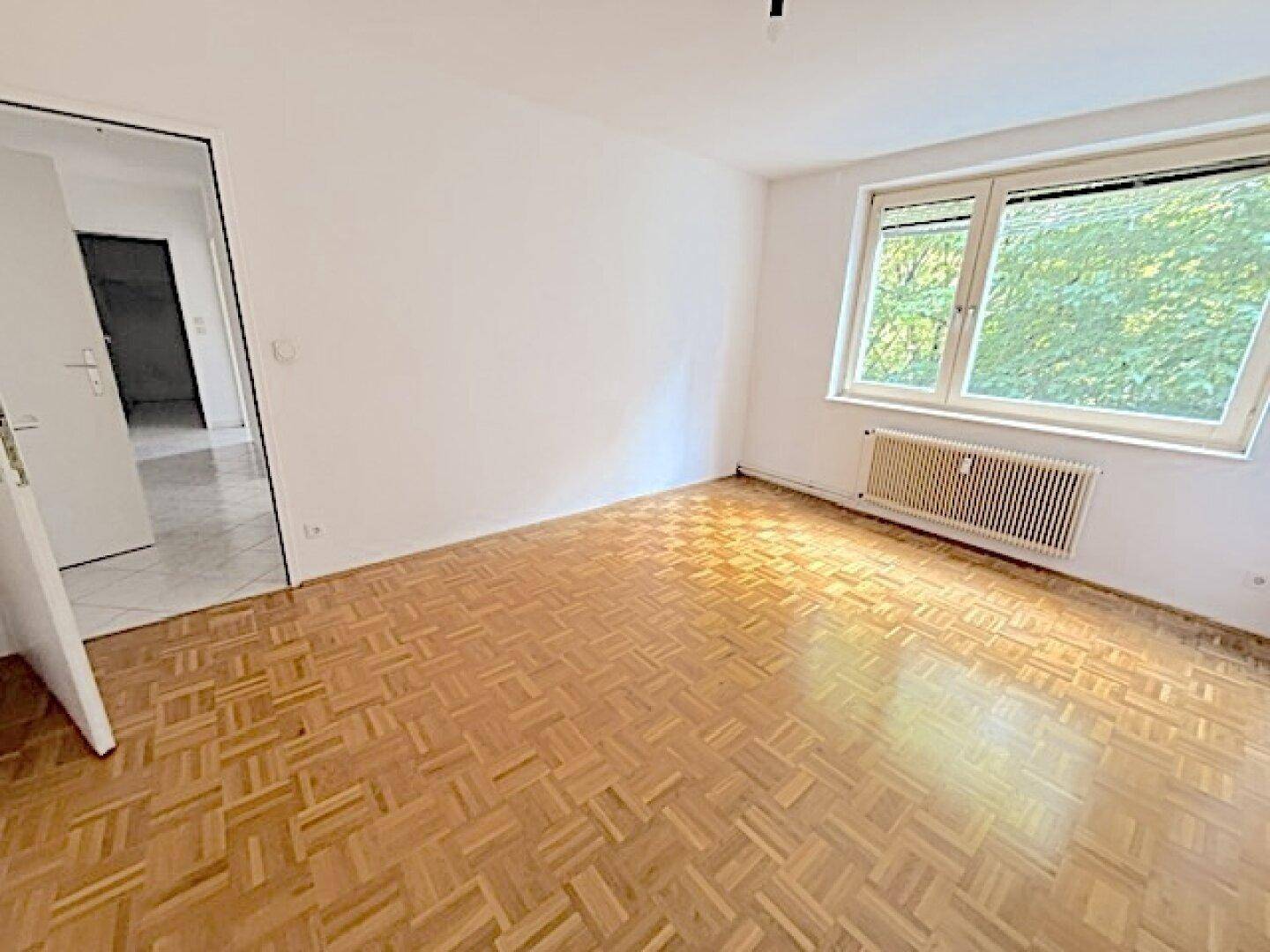 Helles Zimmer mit Fischgrätparkett und Fenster, das den Blick auf die Umgebung freigibt und viel Licht spendet.