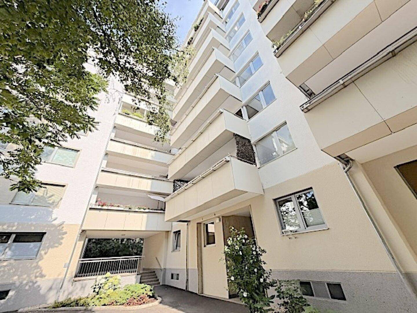 Modernes Wohngebäude mit vielen Balkonen und einer hellen Fassade, umgeben von Bäumen.