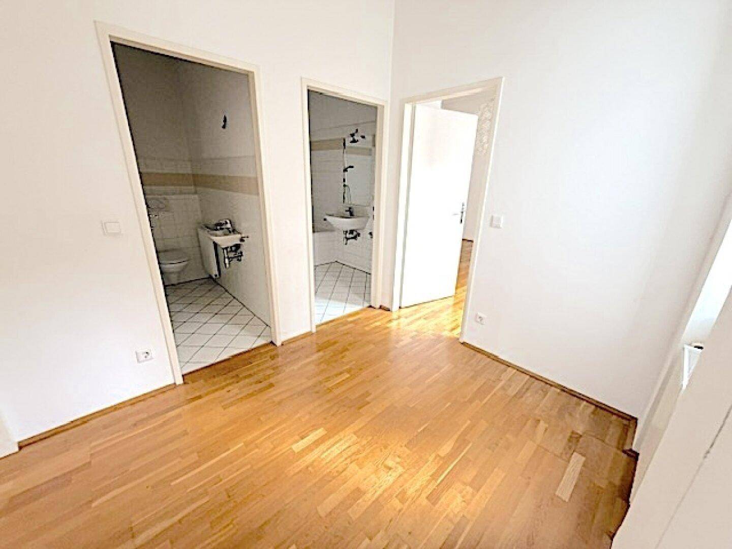 Leerer Raum mit Parkettboden und Zugängen zu zwei gefliesten Sanitärbereichen.