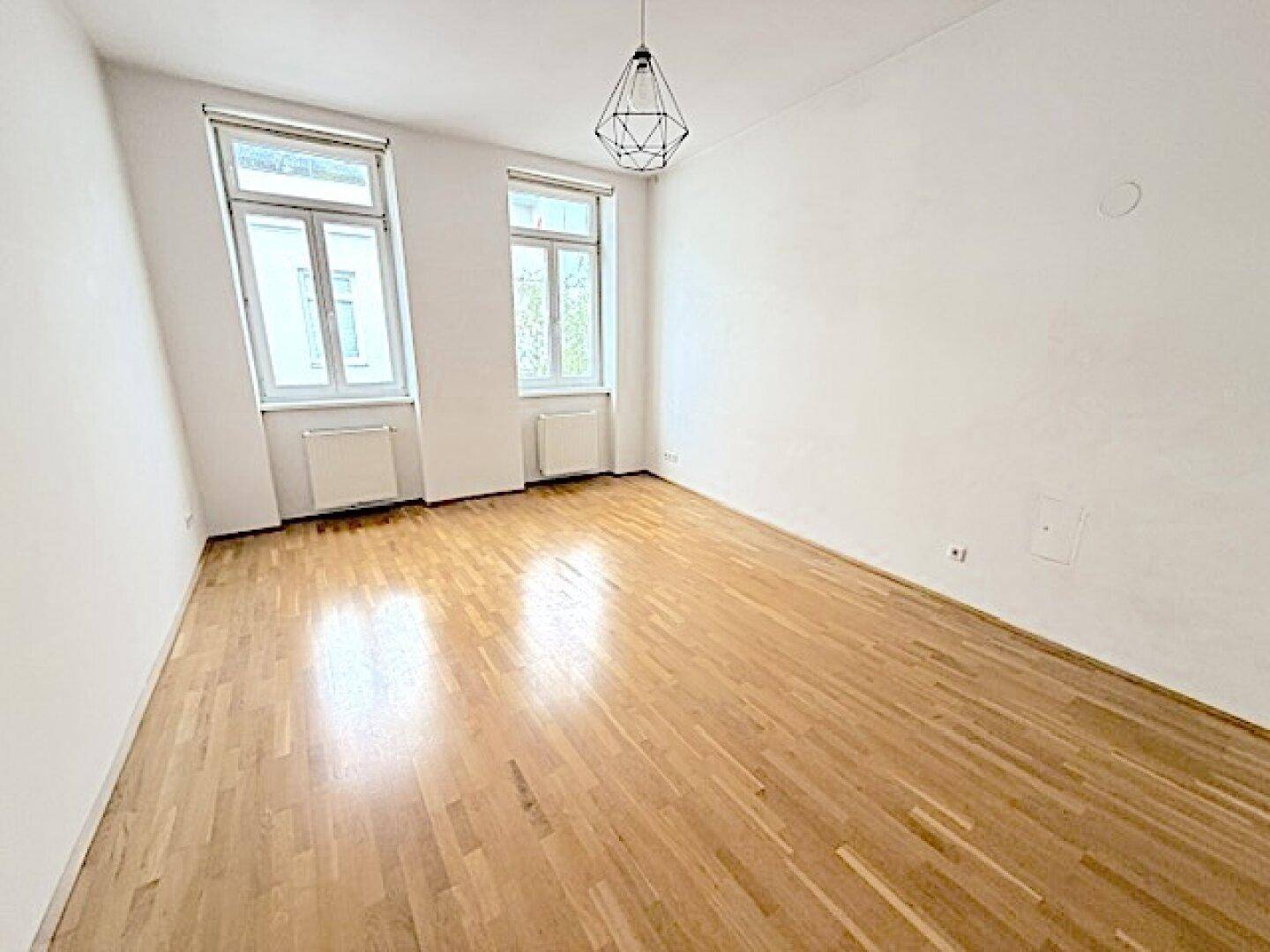 Geräumiges, leeres Zimmer mit hellem Parkettboden und zwei großen Fenstern.